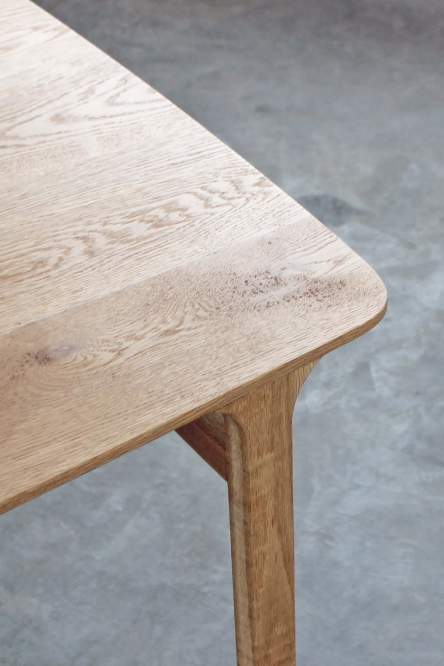 Close-up of the Feynman table's rounded corner in light oak, with a smooth leg flowing up into the top, on a grey concrete floor.