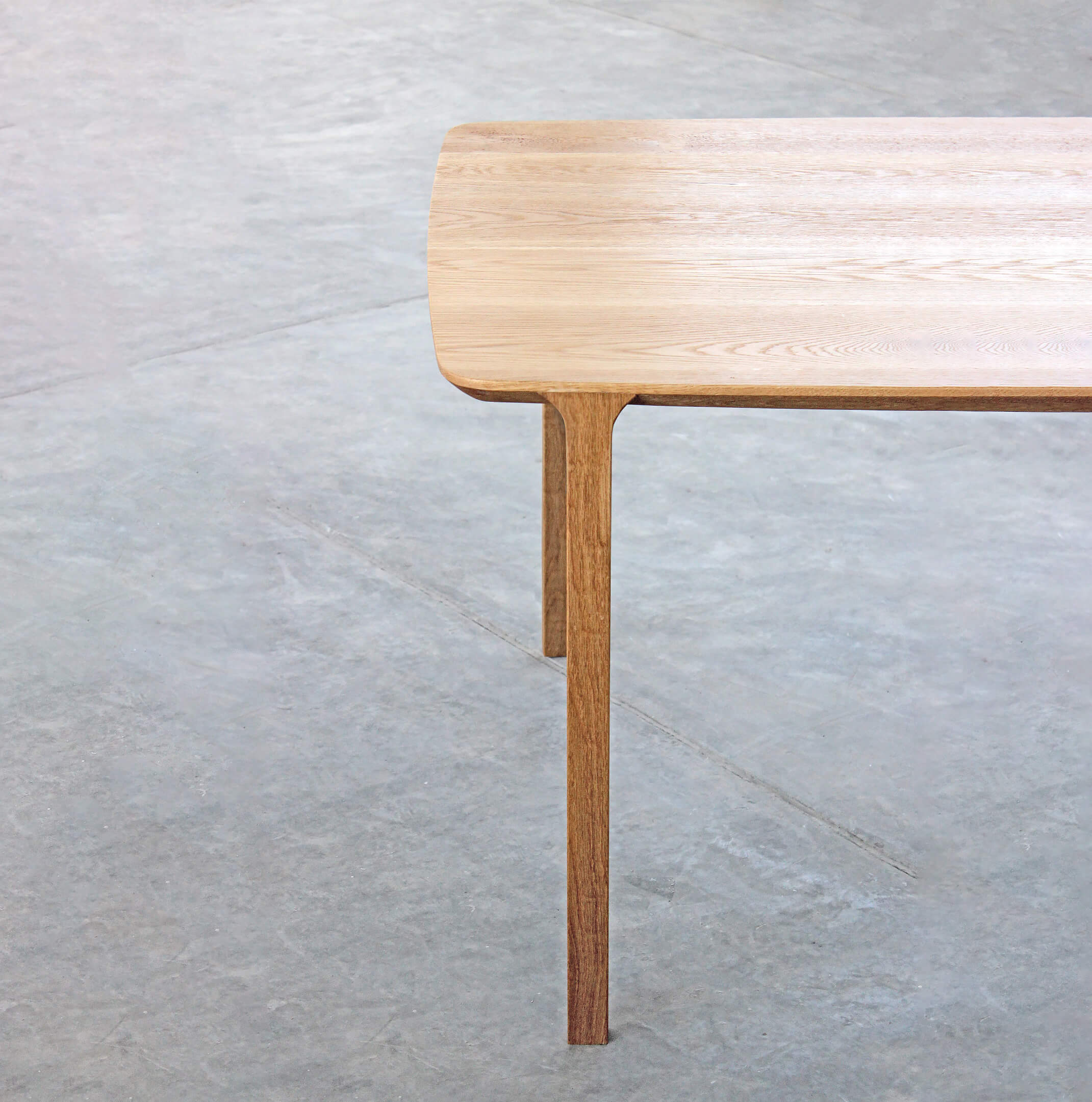 Corner of the light oak Feynman table with one slender, tapered leg rising to the rounded top, on a grey concrete floor.