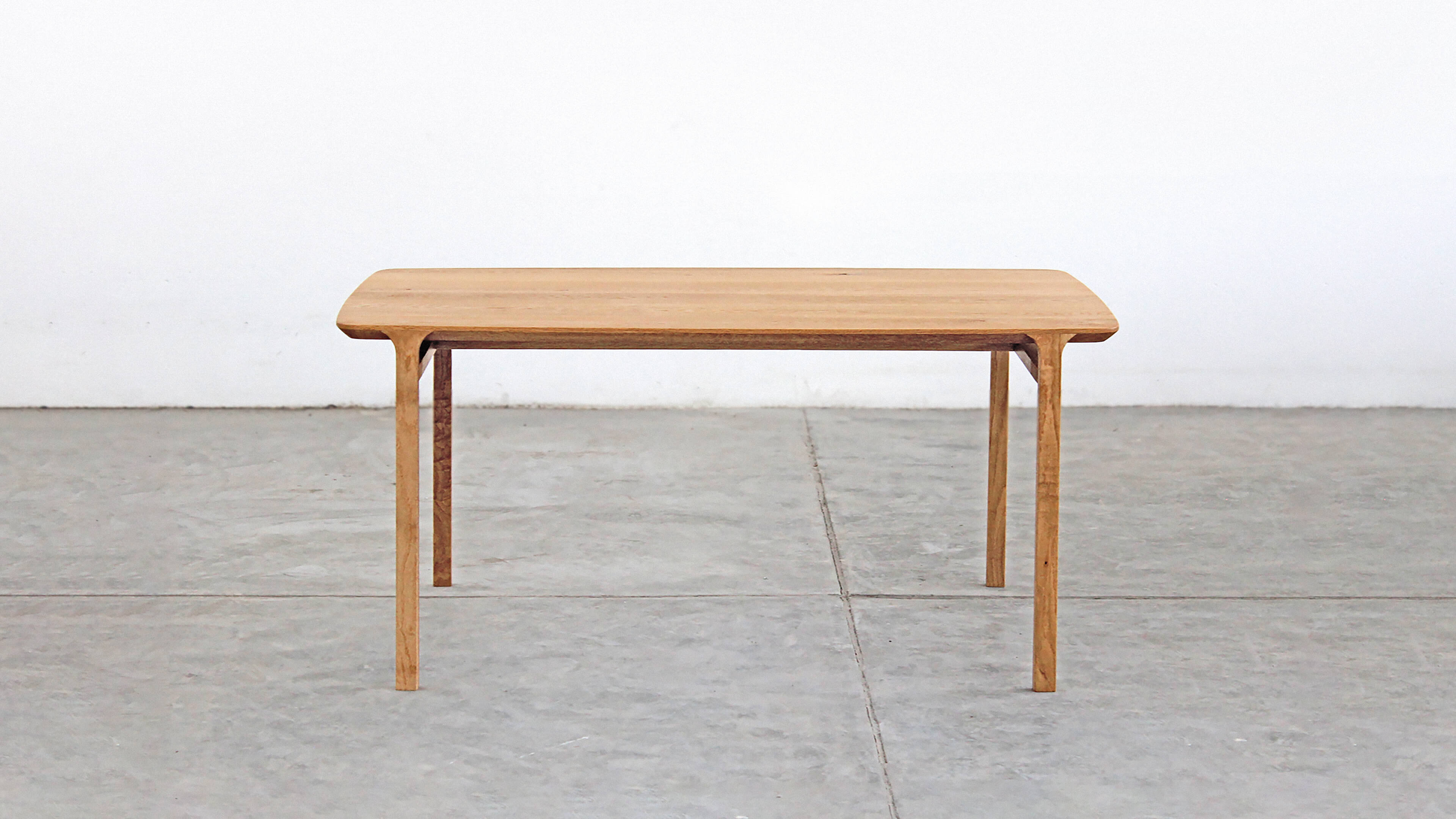 Front view of the whole table: a light oak top with softly rounded corners on four slender legs, against a white wall and a concrete floor.