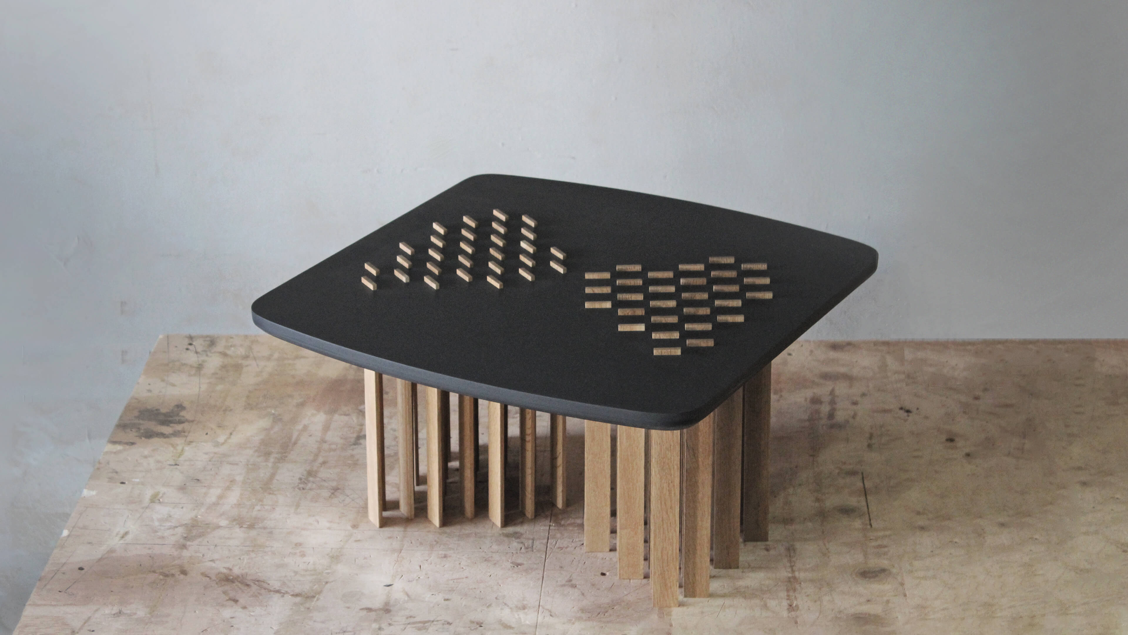 Low Bohr table seen from above, with a black rounded-square top, two groups of small oak pieces inlaid in a pattern, and oak slat legs below.