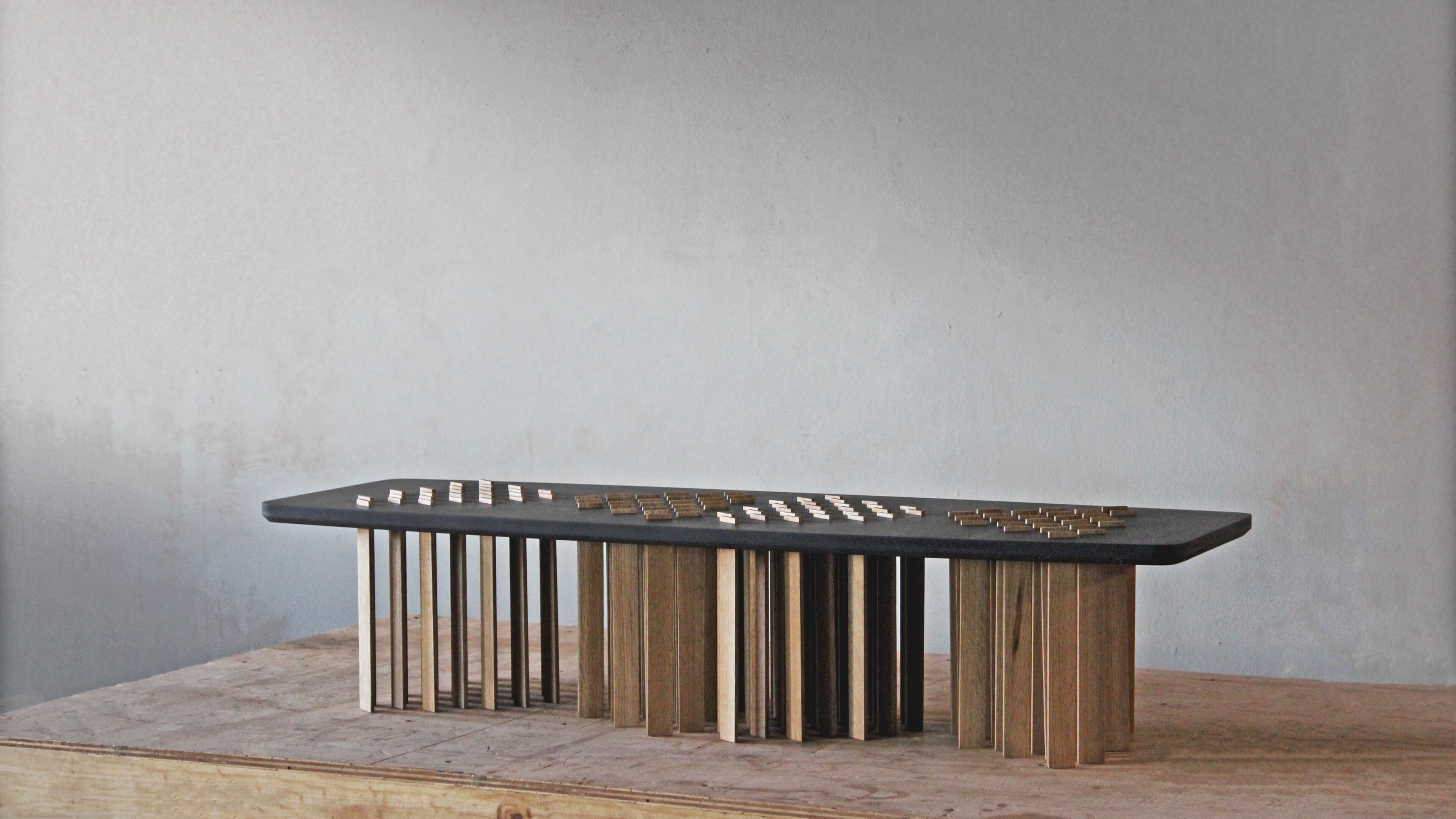 Long, low Bohr table in profile, with a black top carrying three groups of small oak pieces, on a dense row of oak slat legs.