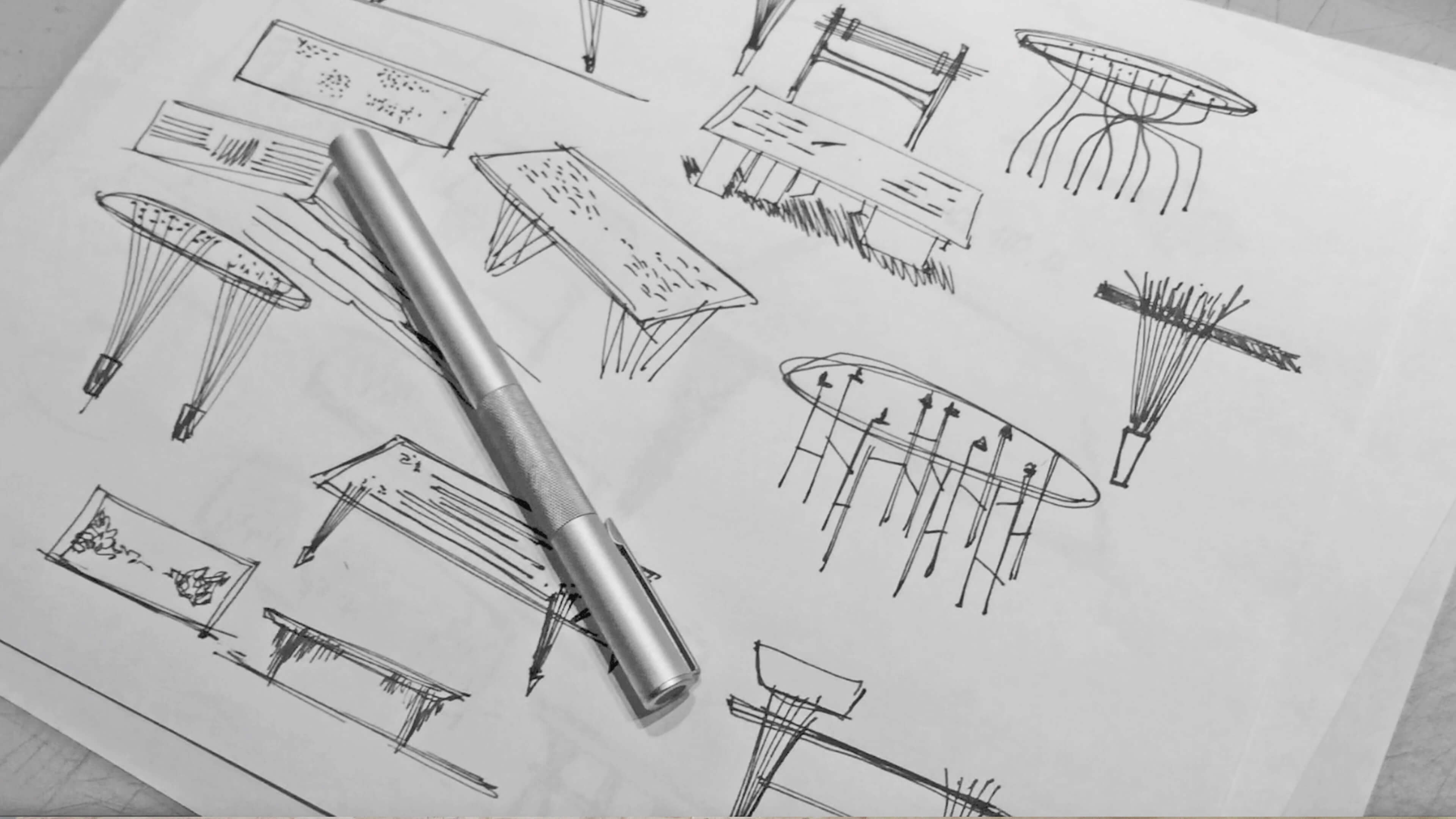 Black and white sheet of ink sketches of tables with slat or rod legs and patterned tops, with a metal pen resting on it.