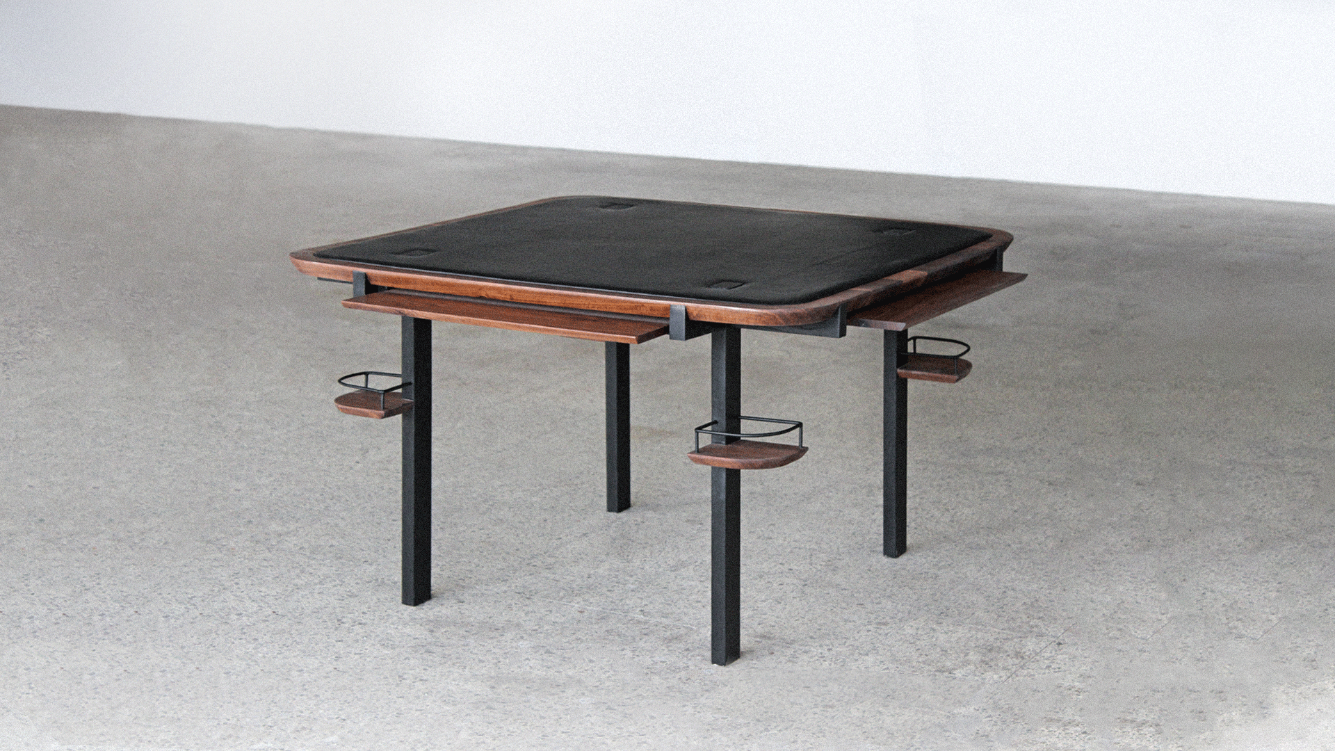 Full Planck game table, with a square black leather top, a walnut edge, black legs, sliding walnut trays and a drinks holder at each corner, on a grey concrete floor.