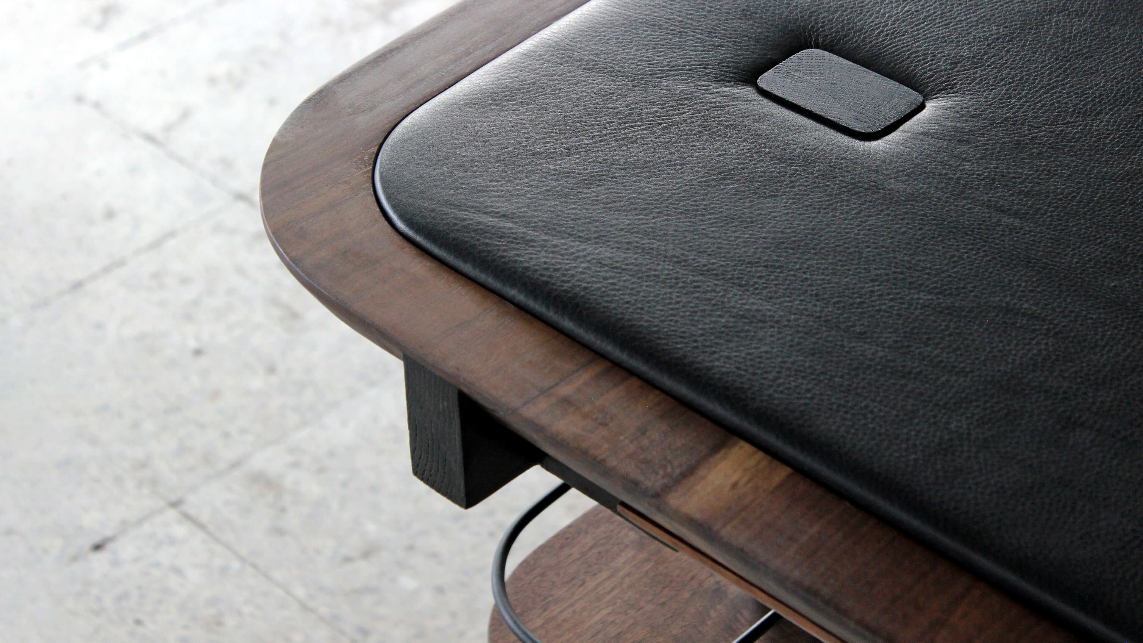 Close-up of the table's rounded corner, with a soft black leather surface, a dark walnut rim and a small square button, above a black wooden support.