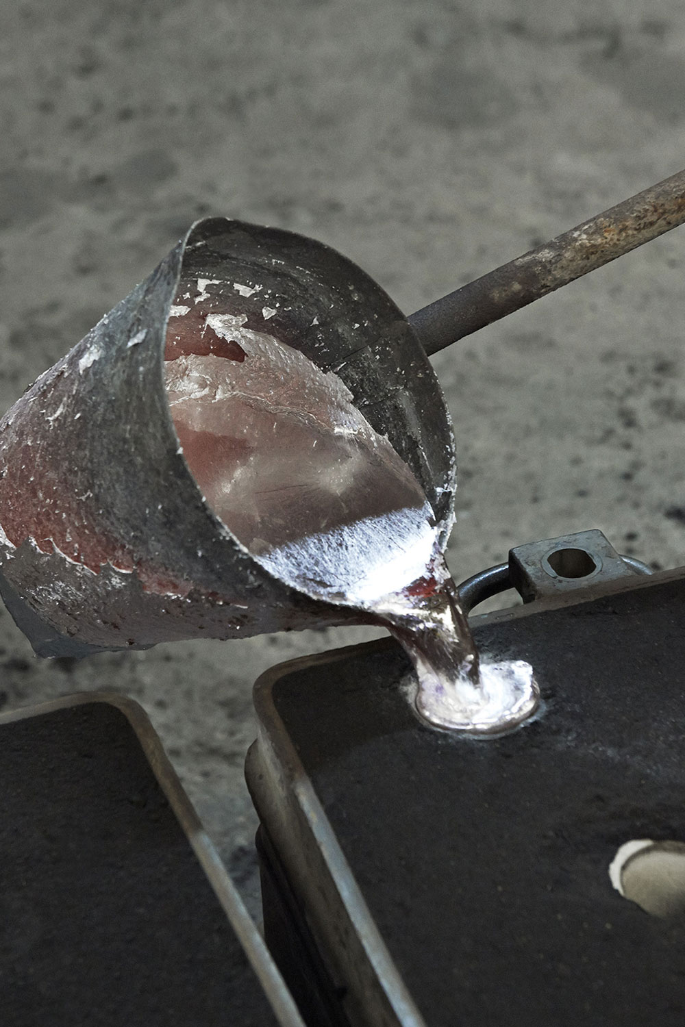 Molten metal poured from a battered ladle into a dark mould in a foundry, with a bright stream landing in the opening.