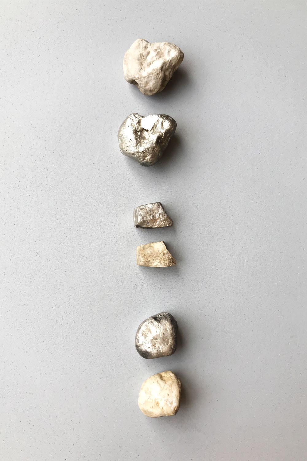 Six stones in a vertical line on a grey surface, rough pale rocks alternating with silver metal versions of them.