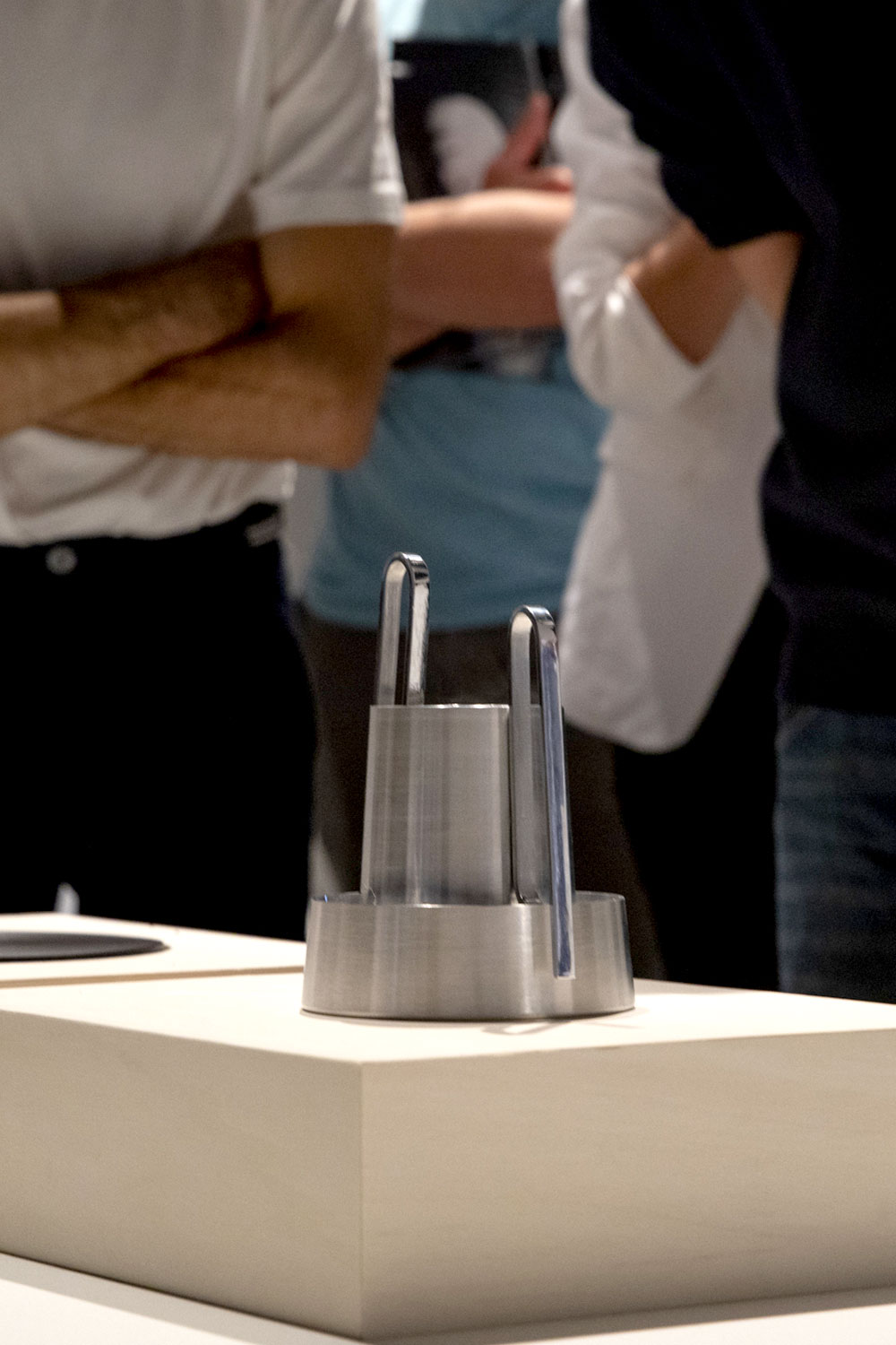 Bloom on a pale wooden plinth at an exhibition, with visitors standing out of focus behind it.