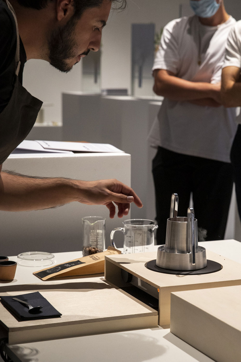 A man in a dark apron leans over a table at an exhibition to demonstrate Bloom, beside a glass jug, a beaker of coffee beans and a black disc.