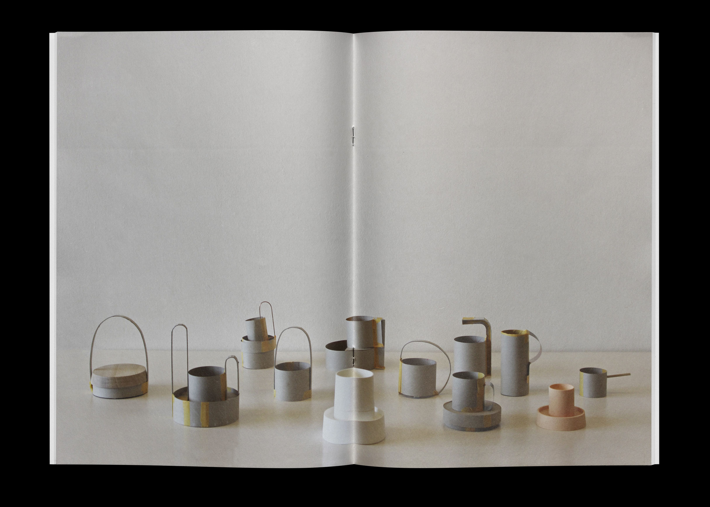 Open booklet spread showing a row of cardboard and paper models of cups, jugs and baskets with handles, in grey, white and pale peach, on a white surface.