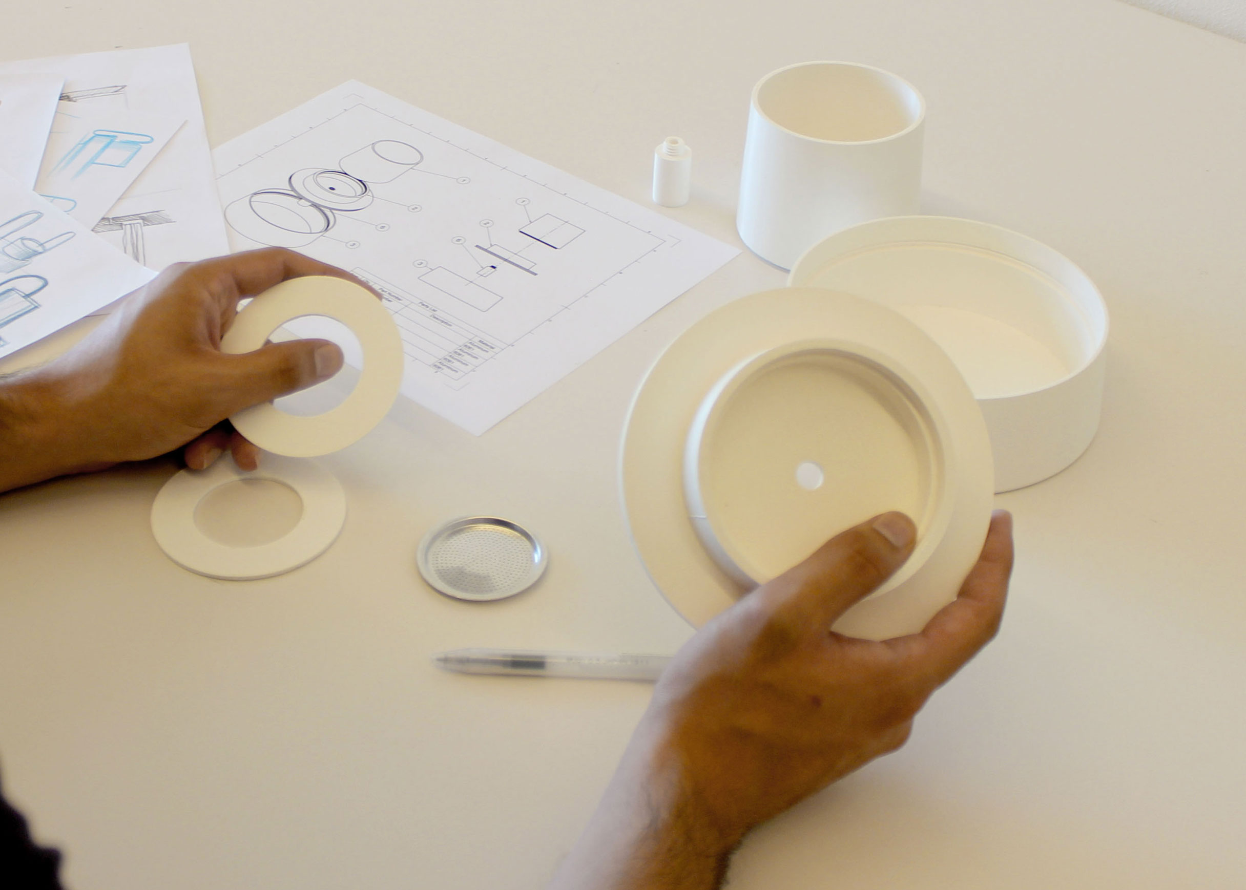 Hands holding white prototype parts, a ring and a conical base, over a technical drawing and a small metal filter basket.