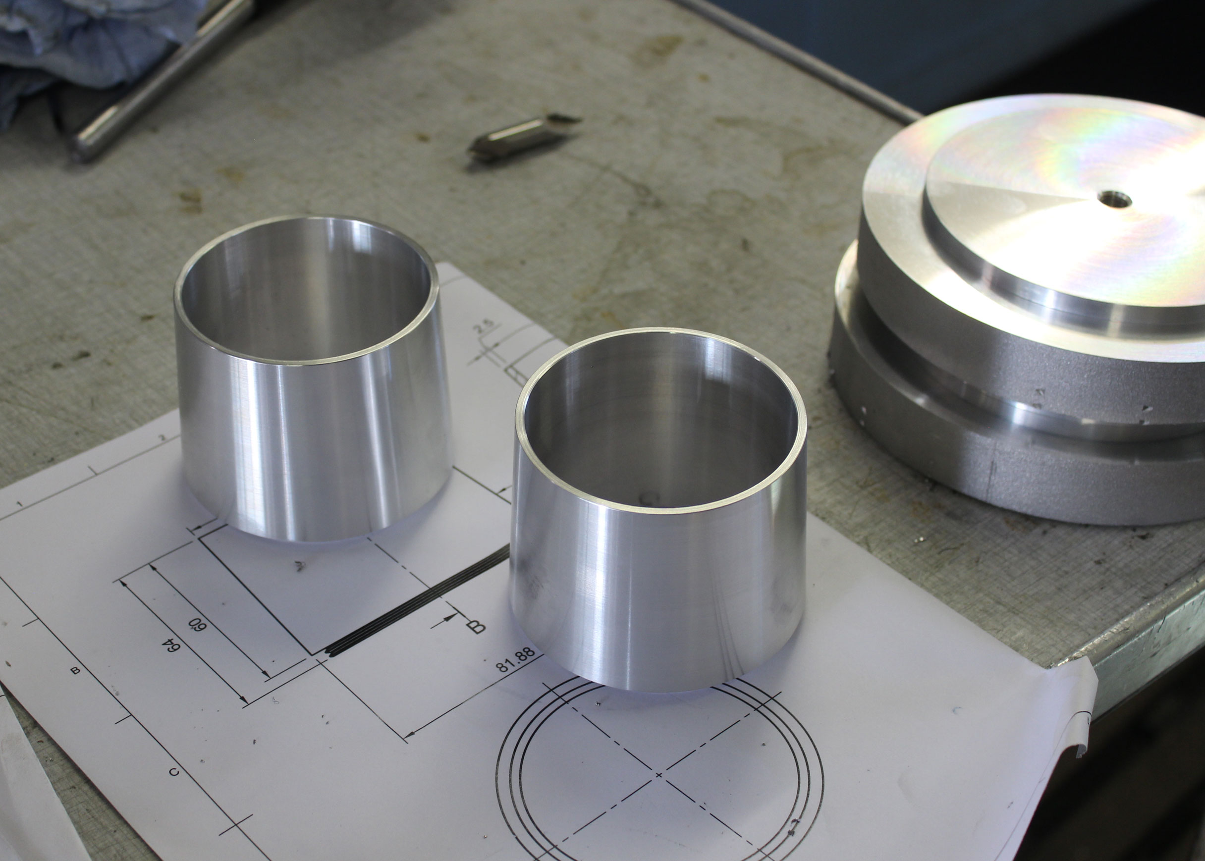 Two polished conical metal parts on a technical drawing in a workshop, beside a larger machined disc with a rainbow sheen.