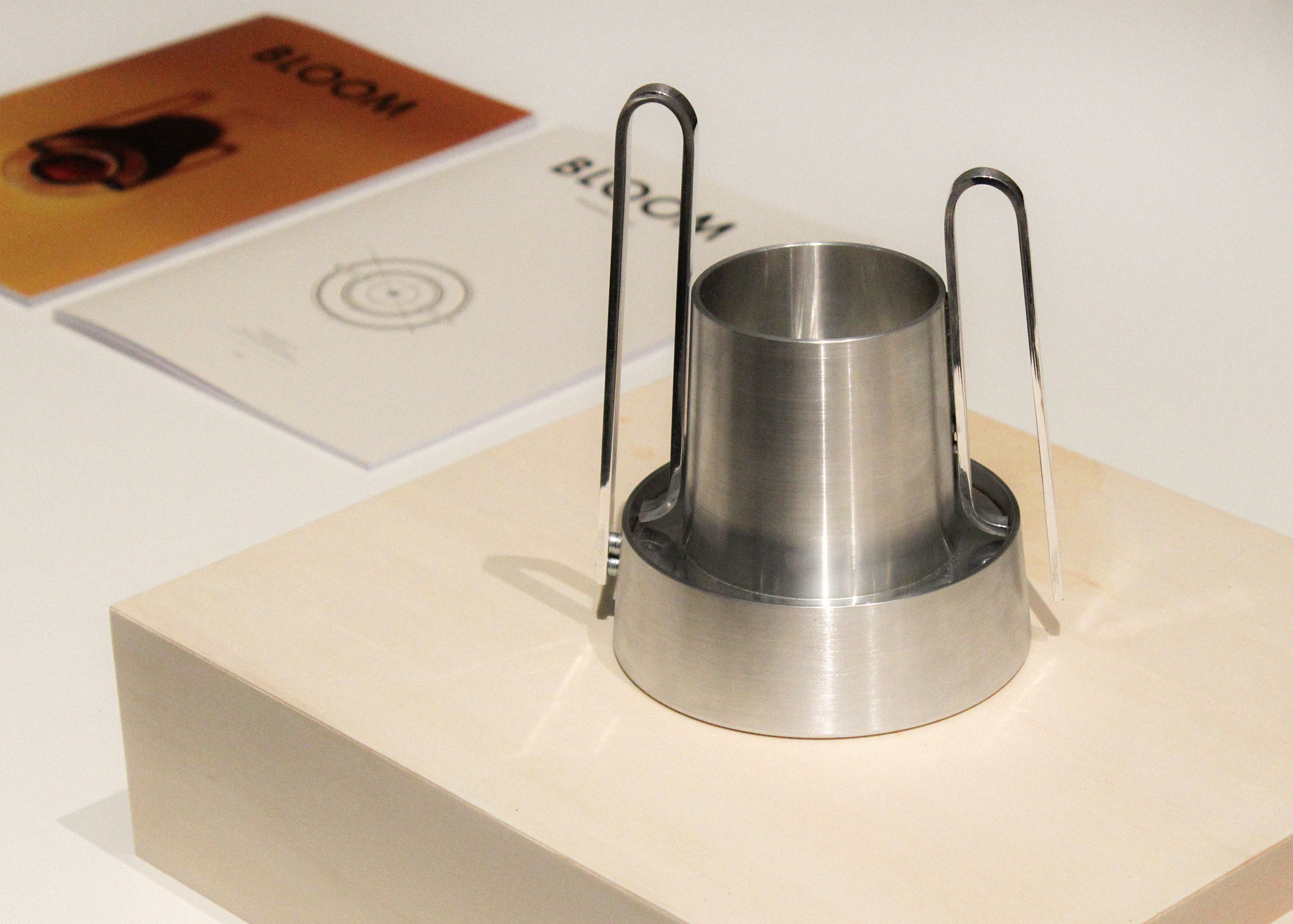 Bloom on a pale wooden plinth, with two Bloom booklets behind it, one orange with a coffee machine on the cover and one white with concentric circles.