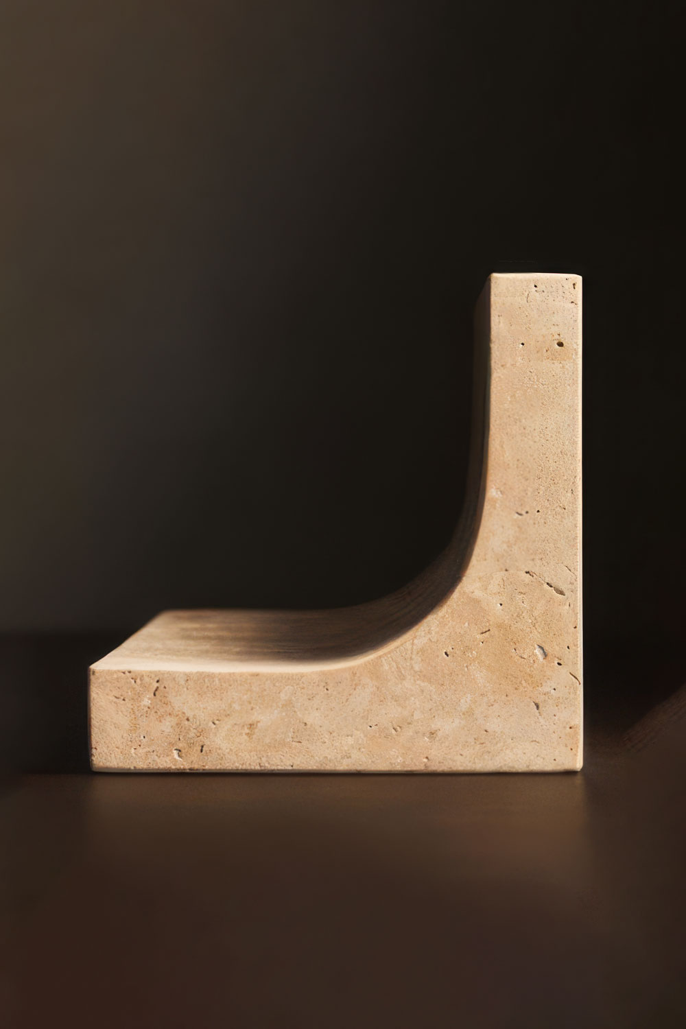 Side profile of the Tondo bookend, an L-shape with a long curved sweep between the upright and the base, in warm cream stone against a dark background.