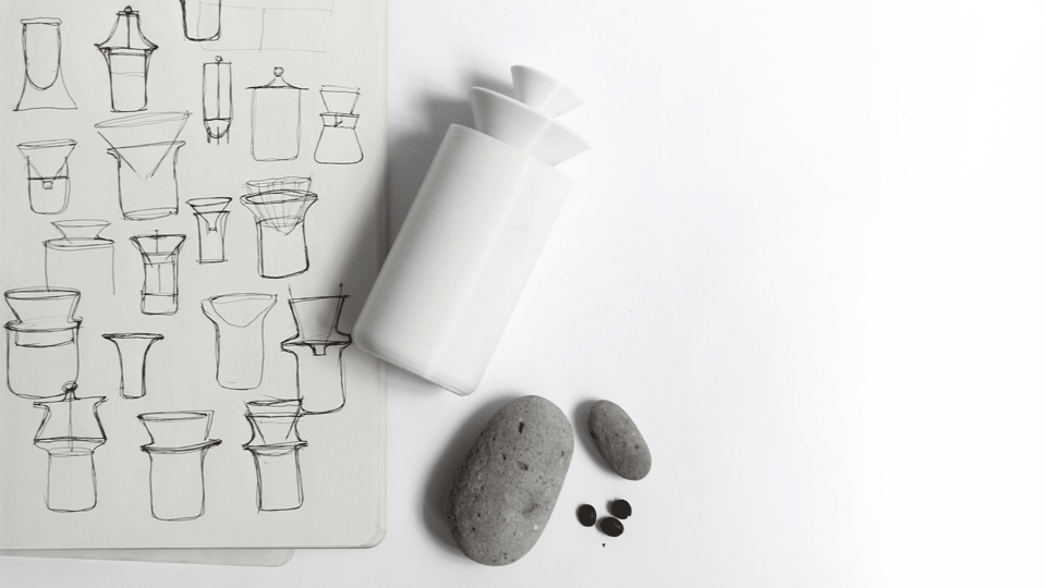 Design process from above: a sketchbook page of dripper sketches, a white model of the cylinder with cone tops, two grey stones and a few coffee beans.