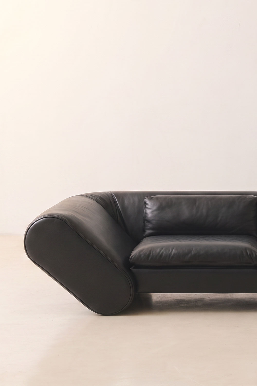 Left half of the black leather Lee sofa, showing its tilted, pill-shaped arm and a single back cushion.
