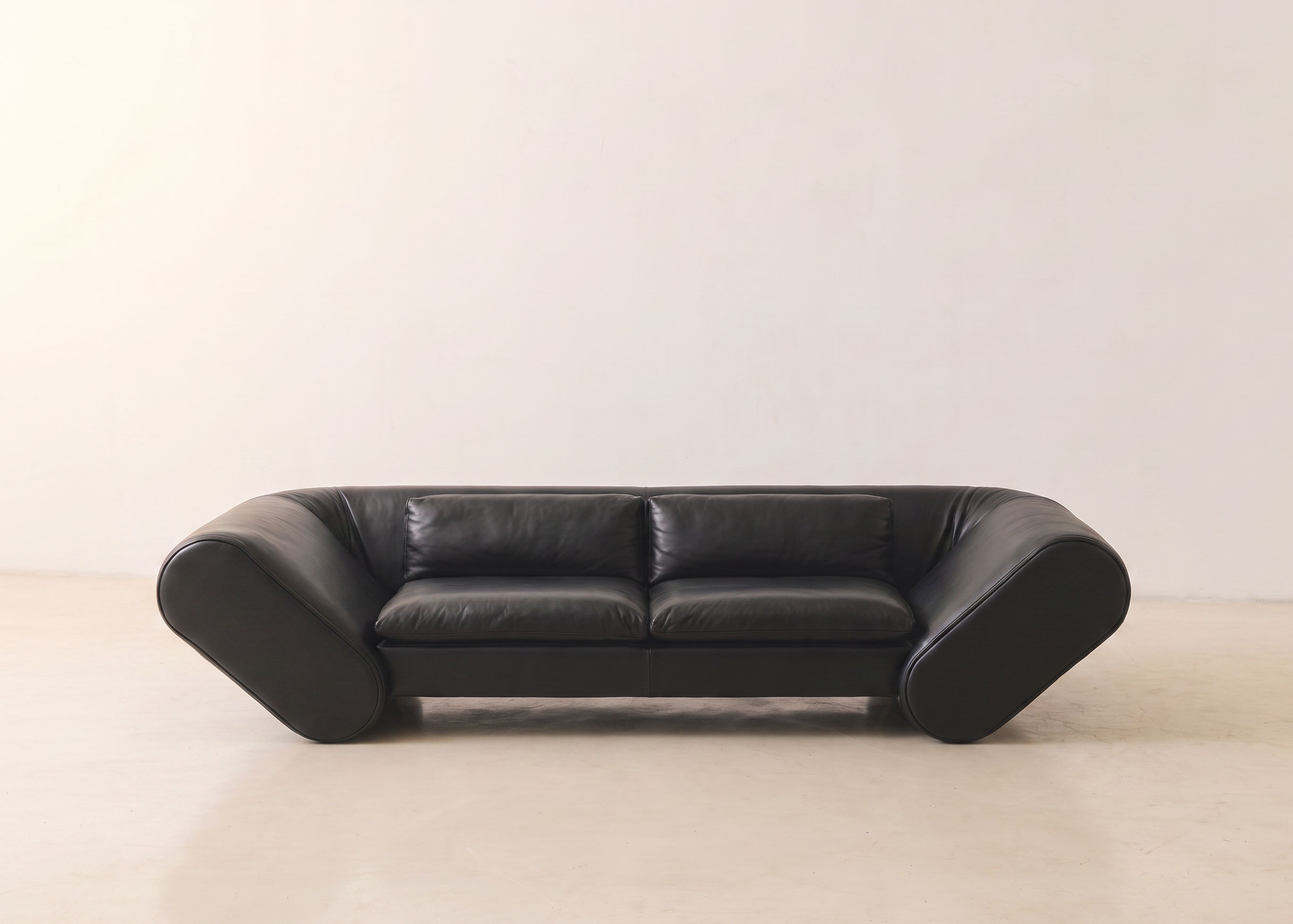 Black leather Lee sofa with two seat cushions and angled pill-shaped arms, in a bright, empty room.