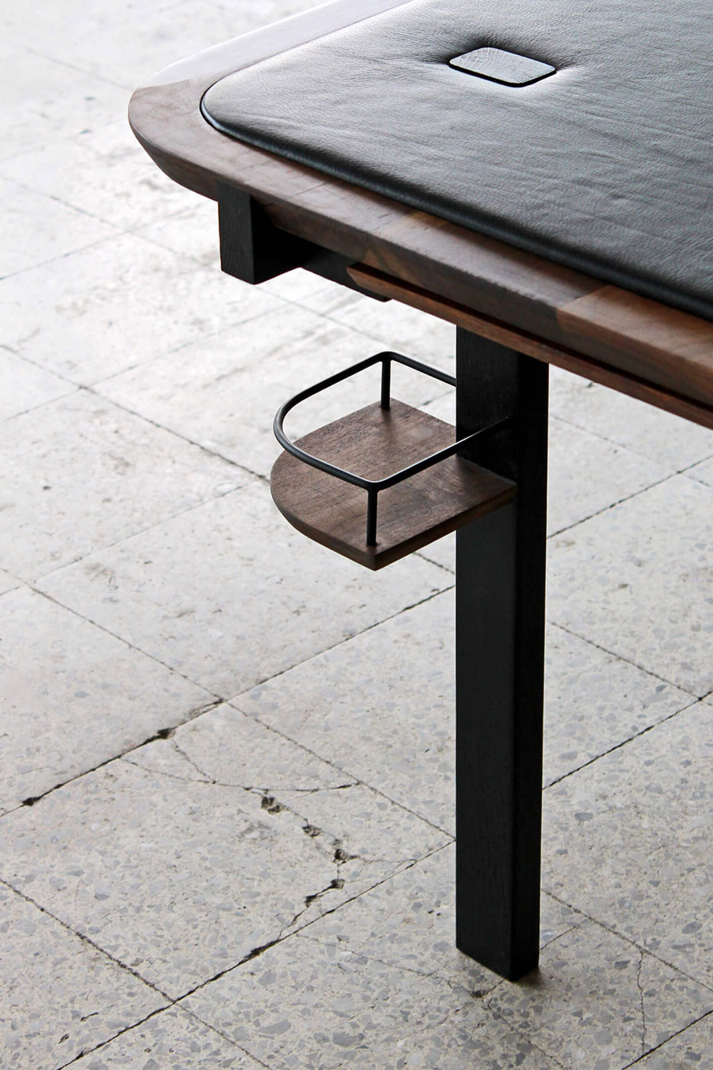 Corner of the Planck game table, with a black leather top and a small square button, a walnut edge, a black leg and a small walnut drinks tray with a black wire rim, on a pale terrazzo floor.