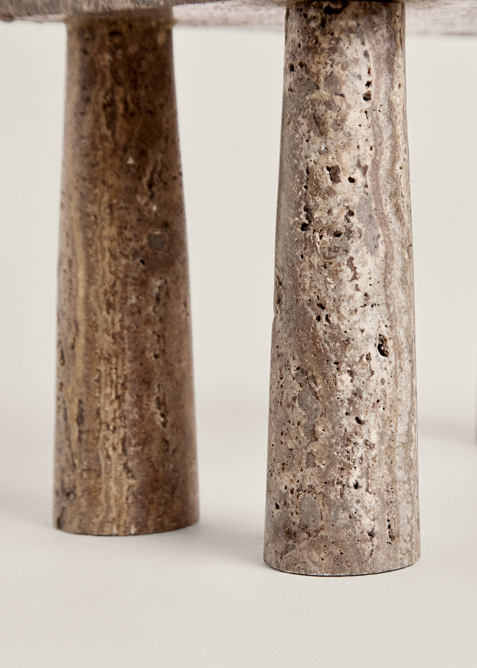 Close-up of two tapered Tap stool legs in veined travertine, one darker and smoother and the other paler and pitted.