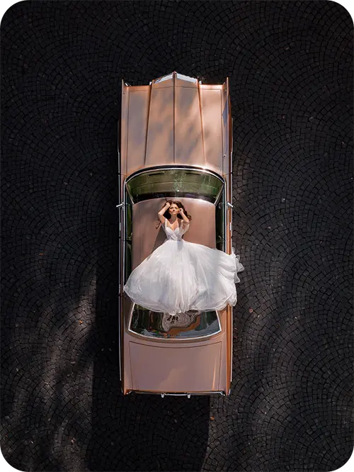Bride in an elegant gown lying across the bonnet of a vintage car, captured from above — romantic aerial view symbolising luxury and timeless style.