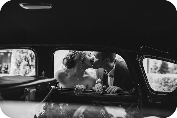 Wedding couple kissing through the window of a vintage car, capturing an intimate moment framed in polished chrome and reflection.