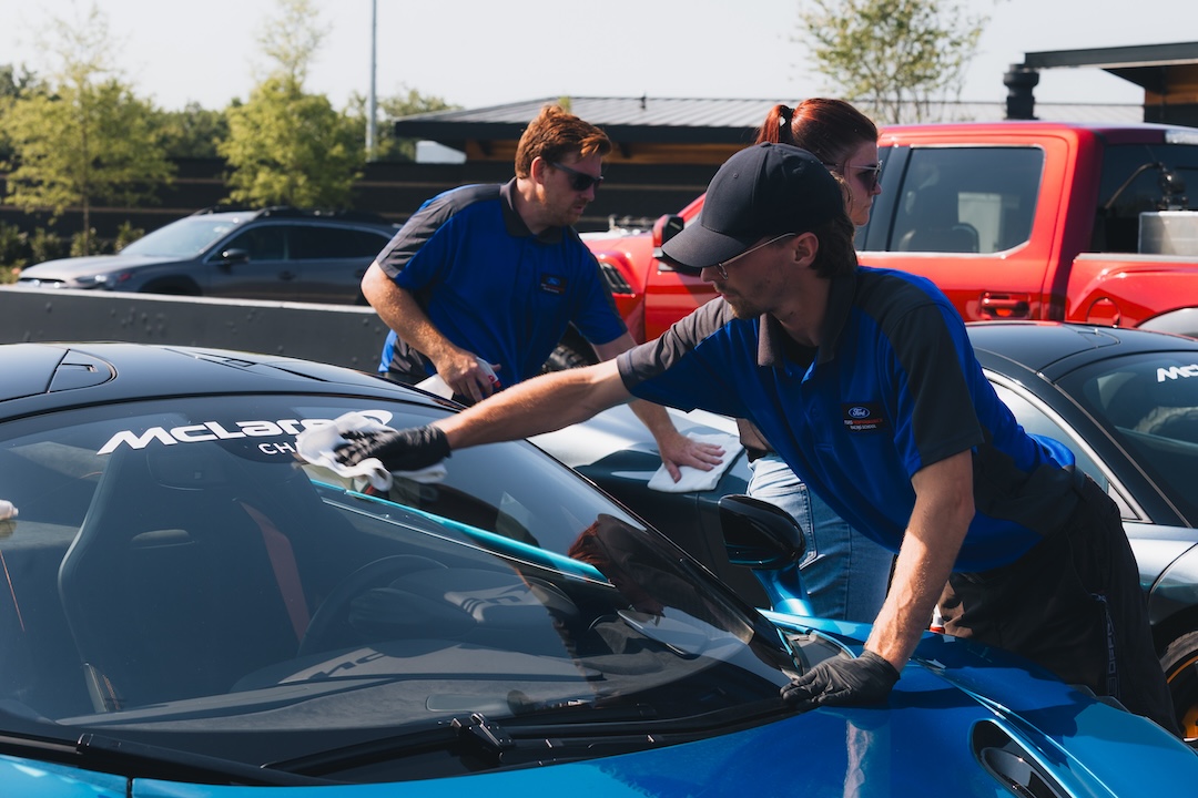 Three people wearing blue shirts cleaning the windshield of a blue McLaren sports car outdoors.