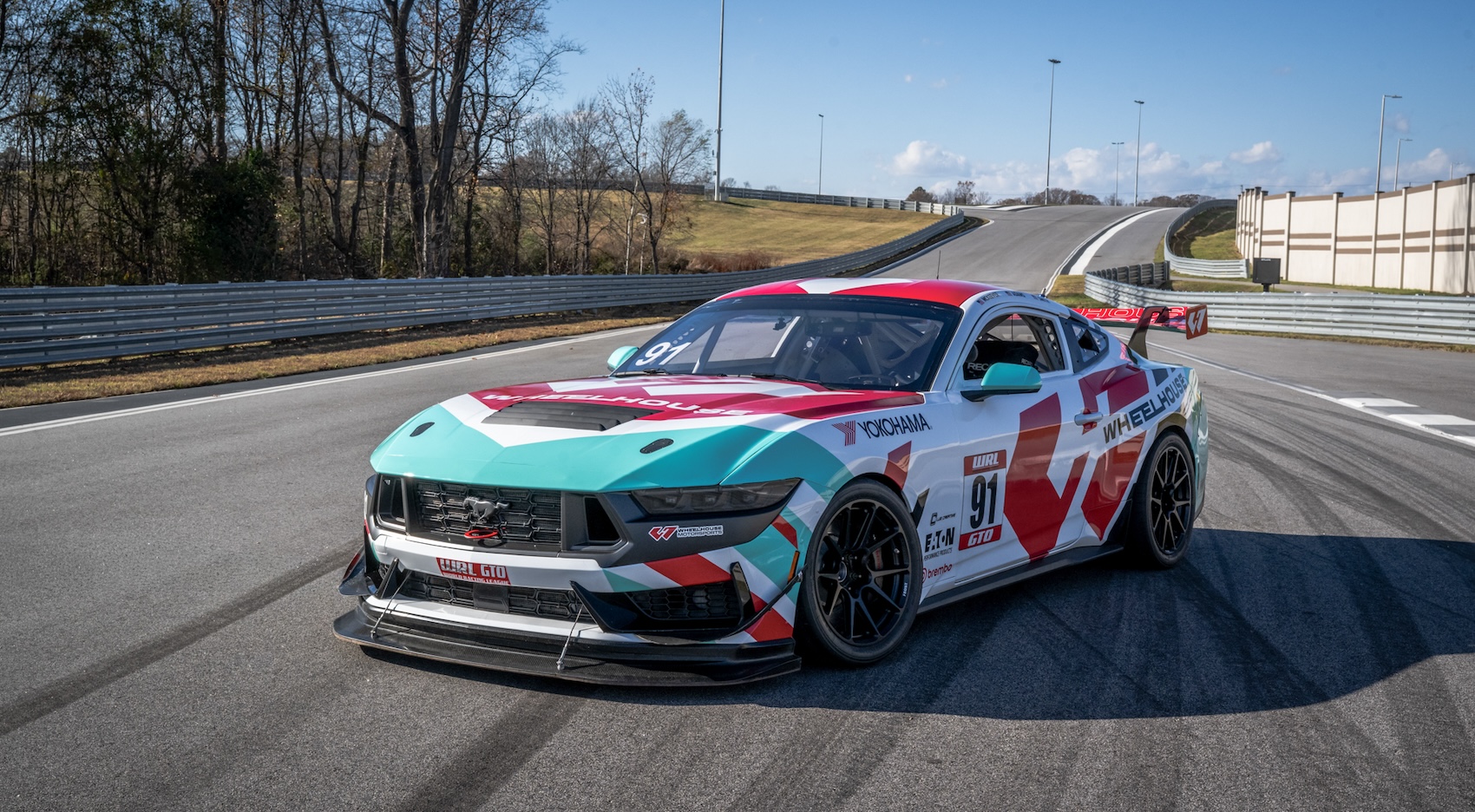 Race car with turquoise, red, and white livery parked on a race track under clear blue sky.
