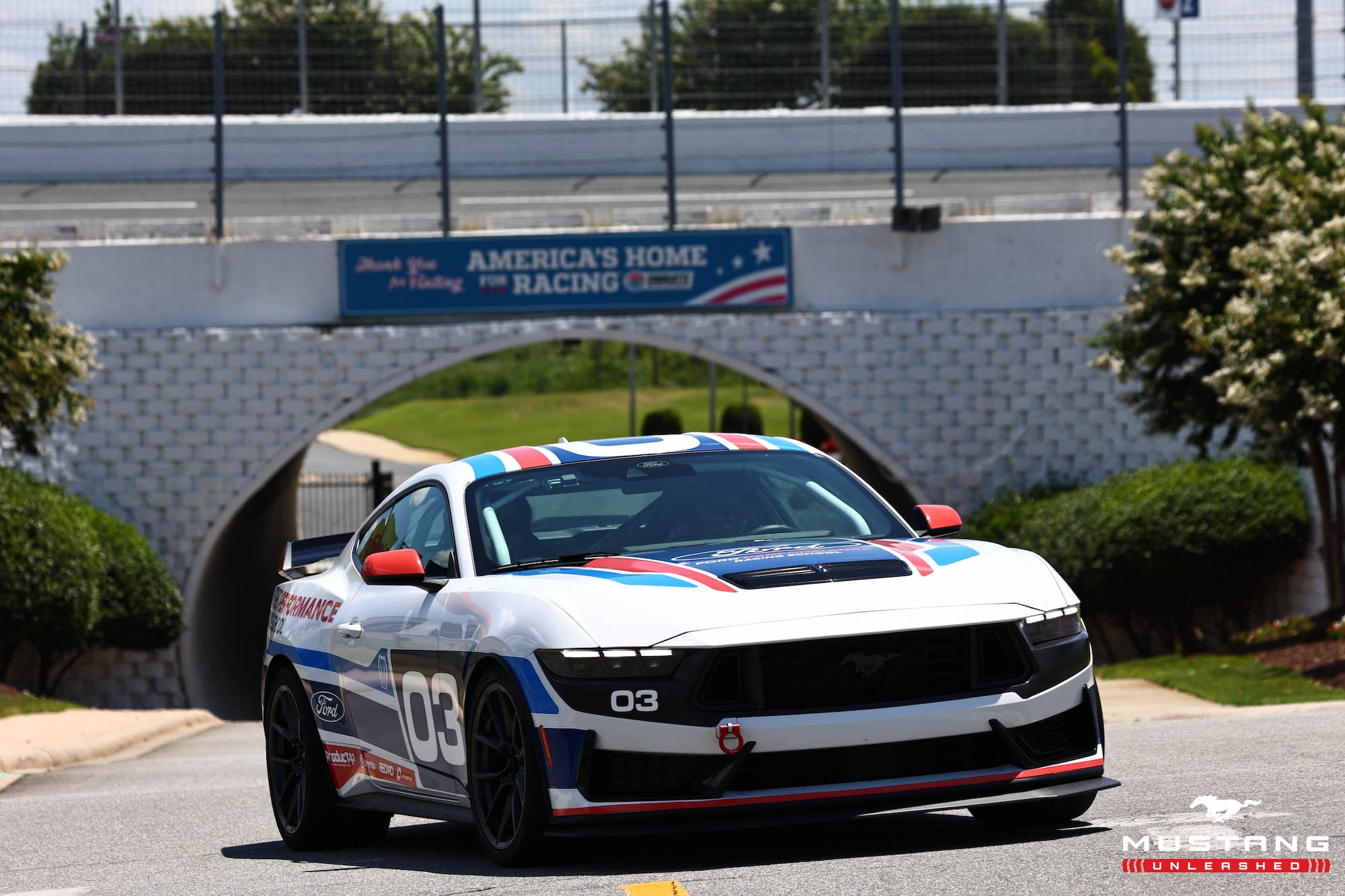 White Ford Mustang race car with blue and red stripes and number 03 on the side, driving near a white brick tunnel with a sign that reads America’s Home for Racing.