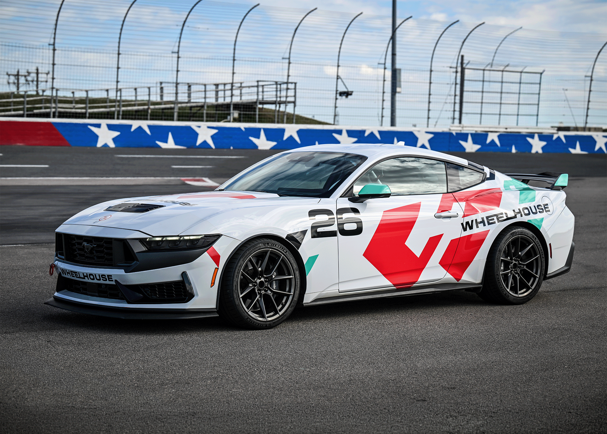 Woman in black jacket and cap speaking to four men standing around a red Mustang car with a water bottle on the rear spoiler.