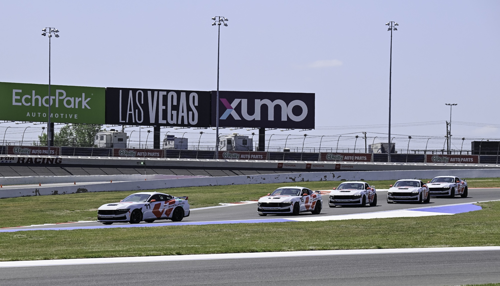 Four people walking near parked red, silver, and blue sports cars, raising their hands in greeting.