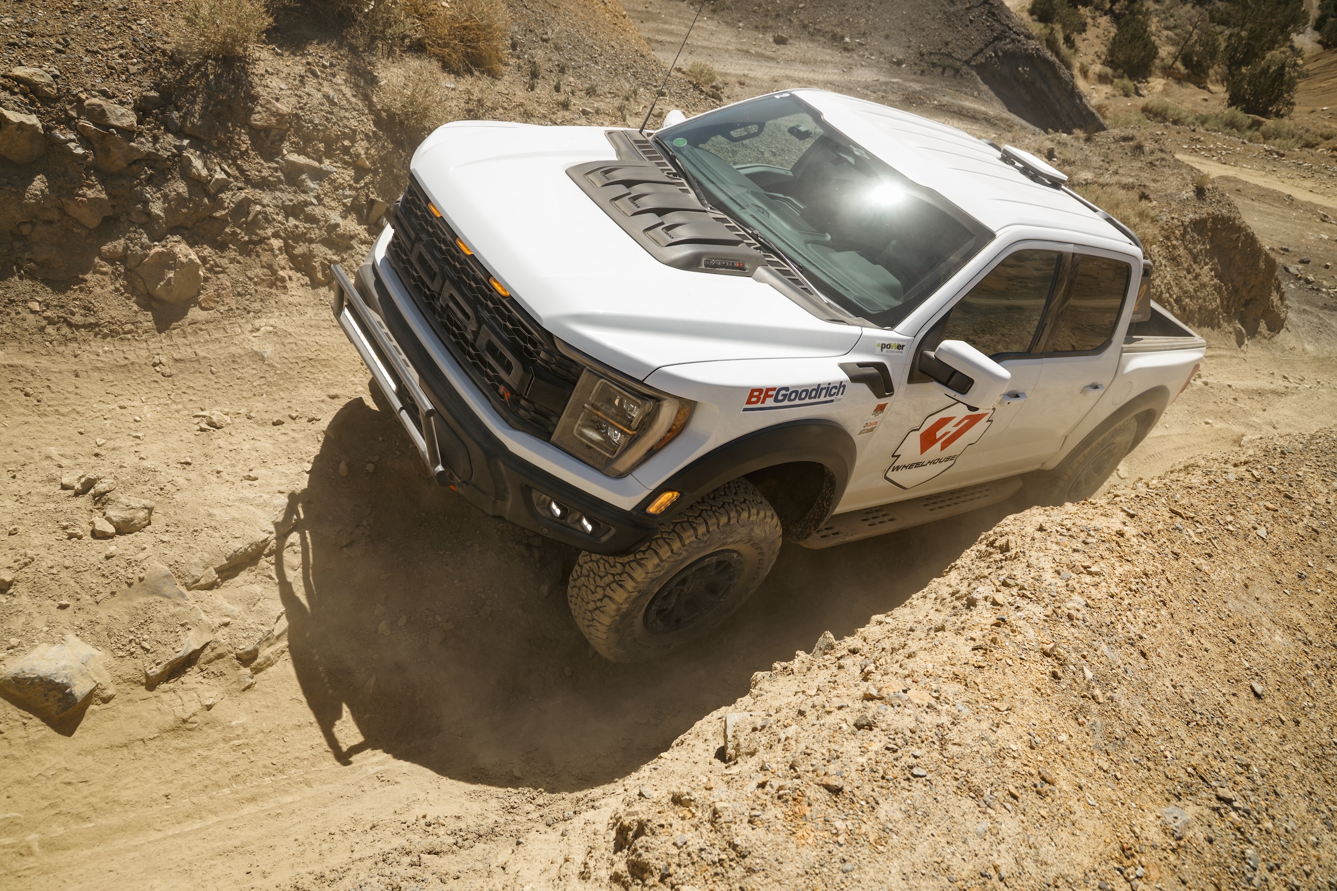 White off-road pickup truck driving on a steep, rocky dirt trail in a desert landscape.