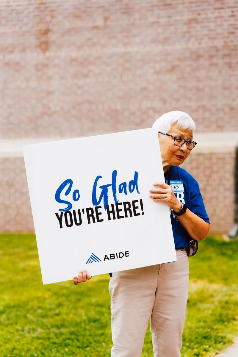 A volunteer holds an Abide sign reading "So glad you're here!" at the Back to School Block Party.