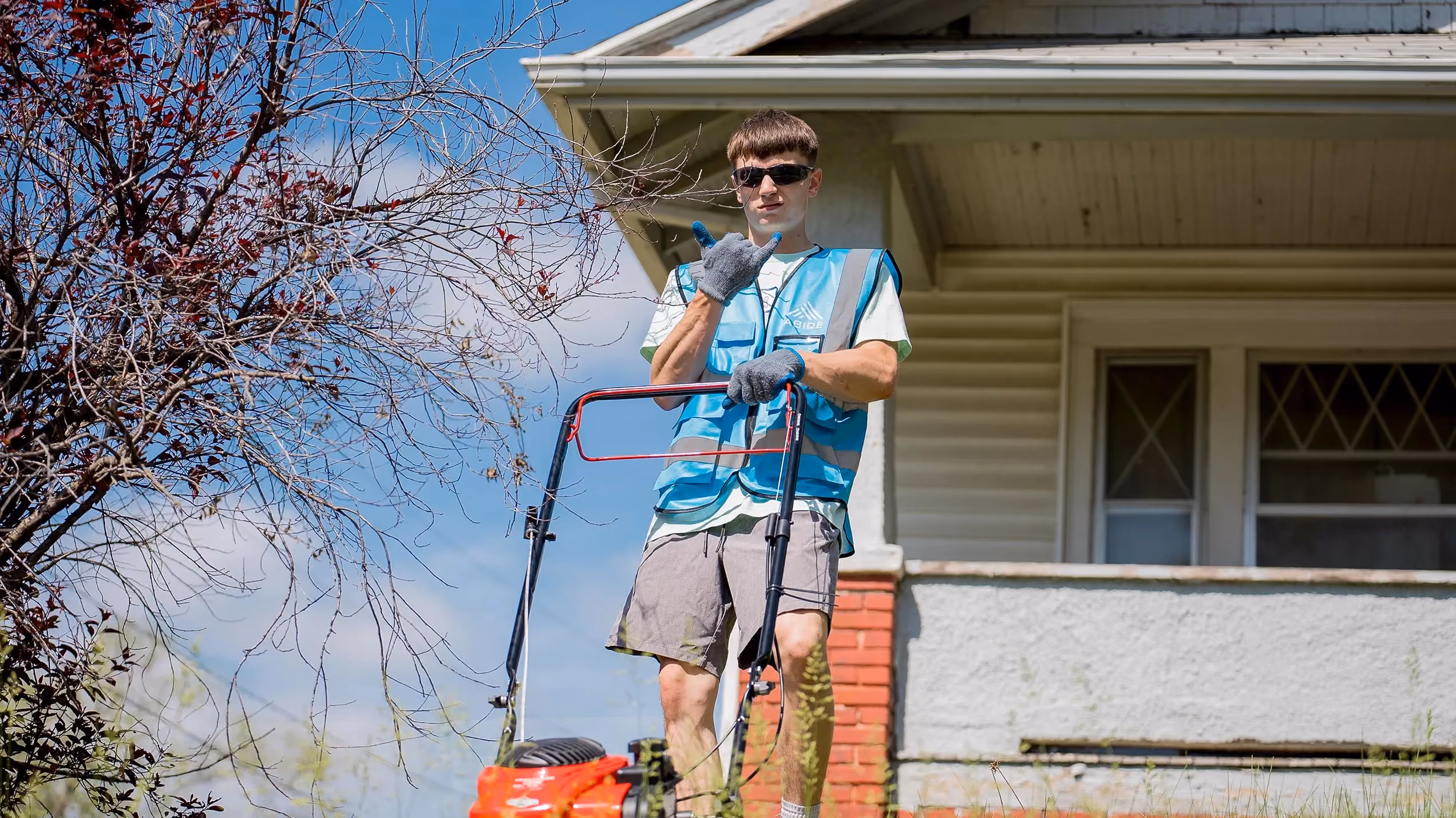 A young volunteer in an Abide vest mows a front yard and gives a friendly wave.