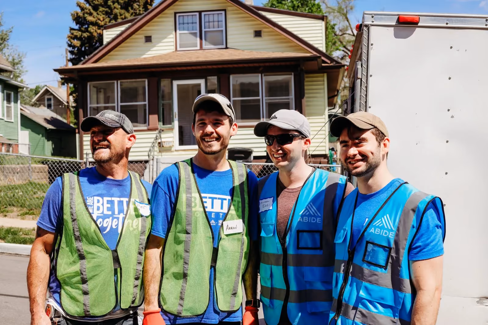 Four volunteers in safety vests and Abide shirts smile in front of a house during Spring Clean Up.