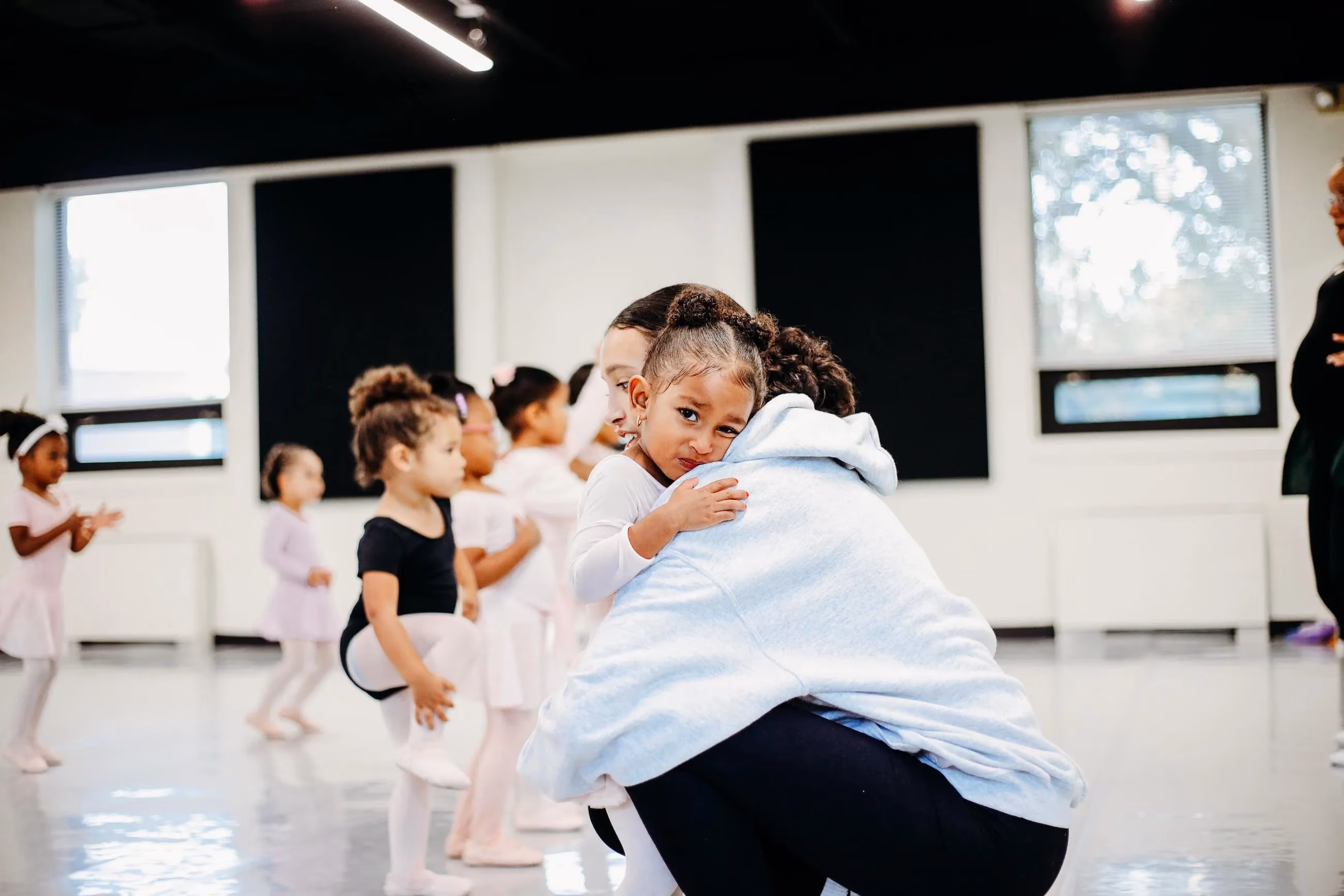 A young dancer hugs her teacher during a ballet class as other kids practice nearby.