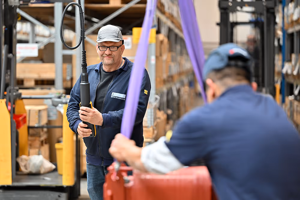 Zwei Männer arbeiten mit Hebeausrüstung in einem Lager voller Kartons.