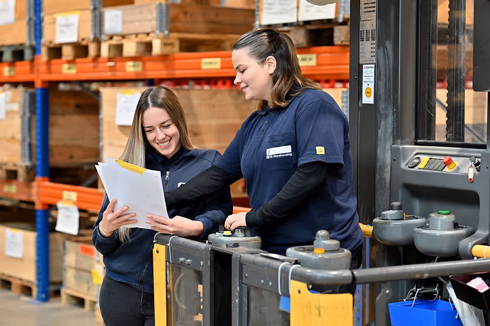 Zwei Frauen in einem Lagerhaus, eine zeigt der anderen lächelnd Dokumente, umgeben von Regalen mit Kartons.