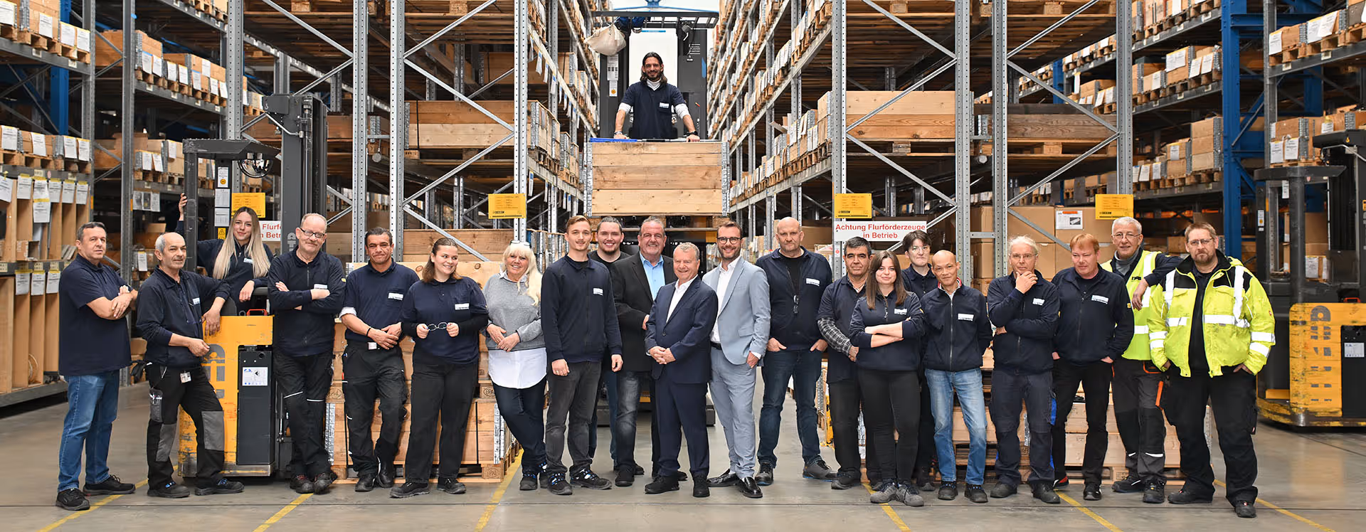 Gruppenfoto von 22 Mitarbeitern in einem Lagerhaus, umgeben von Regalen mit Holzkisten.