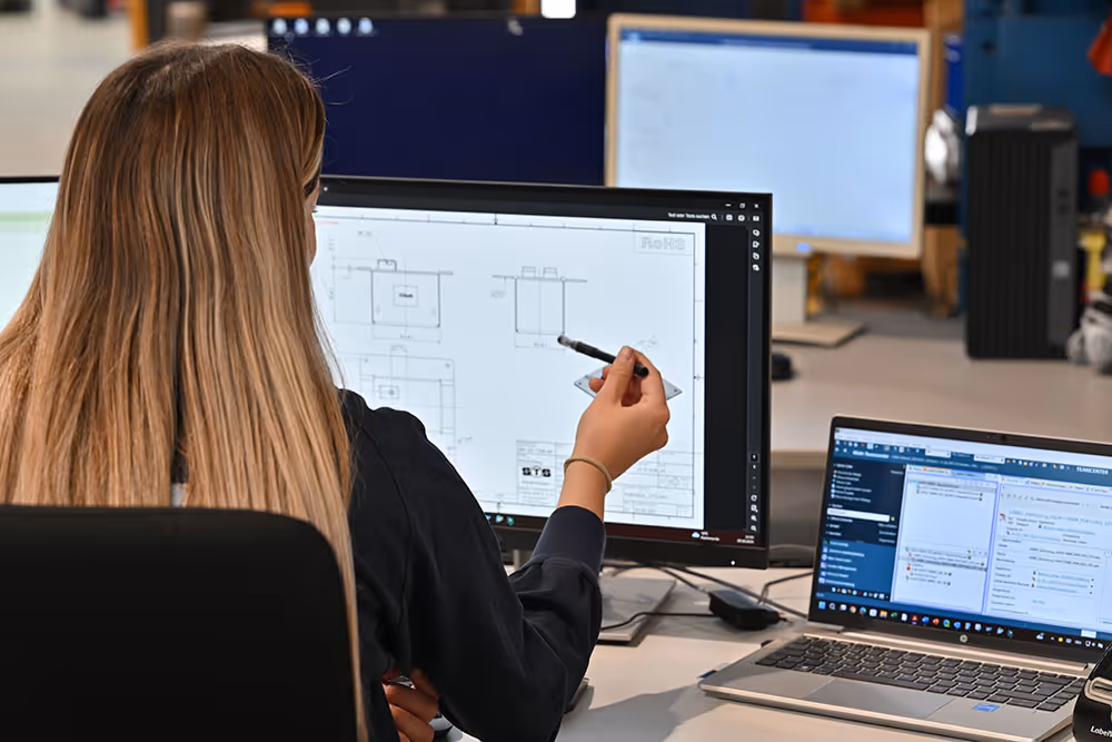 Frau mit langen Haaren, die mit einem Stift auf technische Zeichnungen auf einem Computerbildschirm zeigt, vor einem Laptop mit geöffneten Dokumenten.