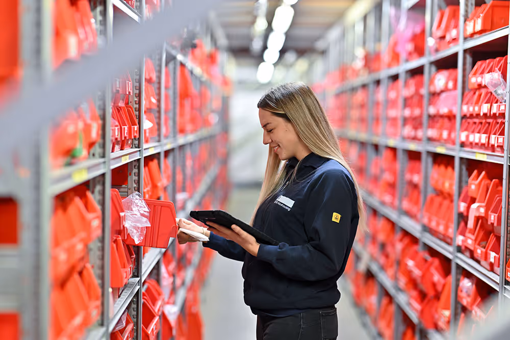 Junge Lagerarbeiterin in dunkelblauem Pullover überprüft rote Behälter im Lager mit Tablet.