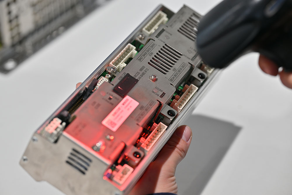 Nahaufnahme einer Hand, die ein elektronisches Gerät mit verschiedenen Anschlüssen hält, während ein Scanner mit rotem Licht darauf gerichtet ist.