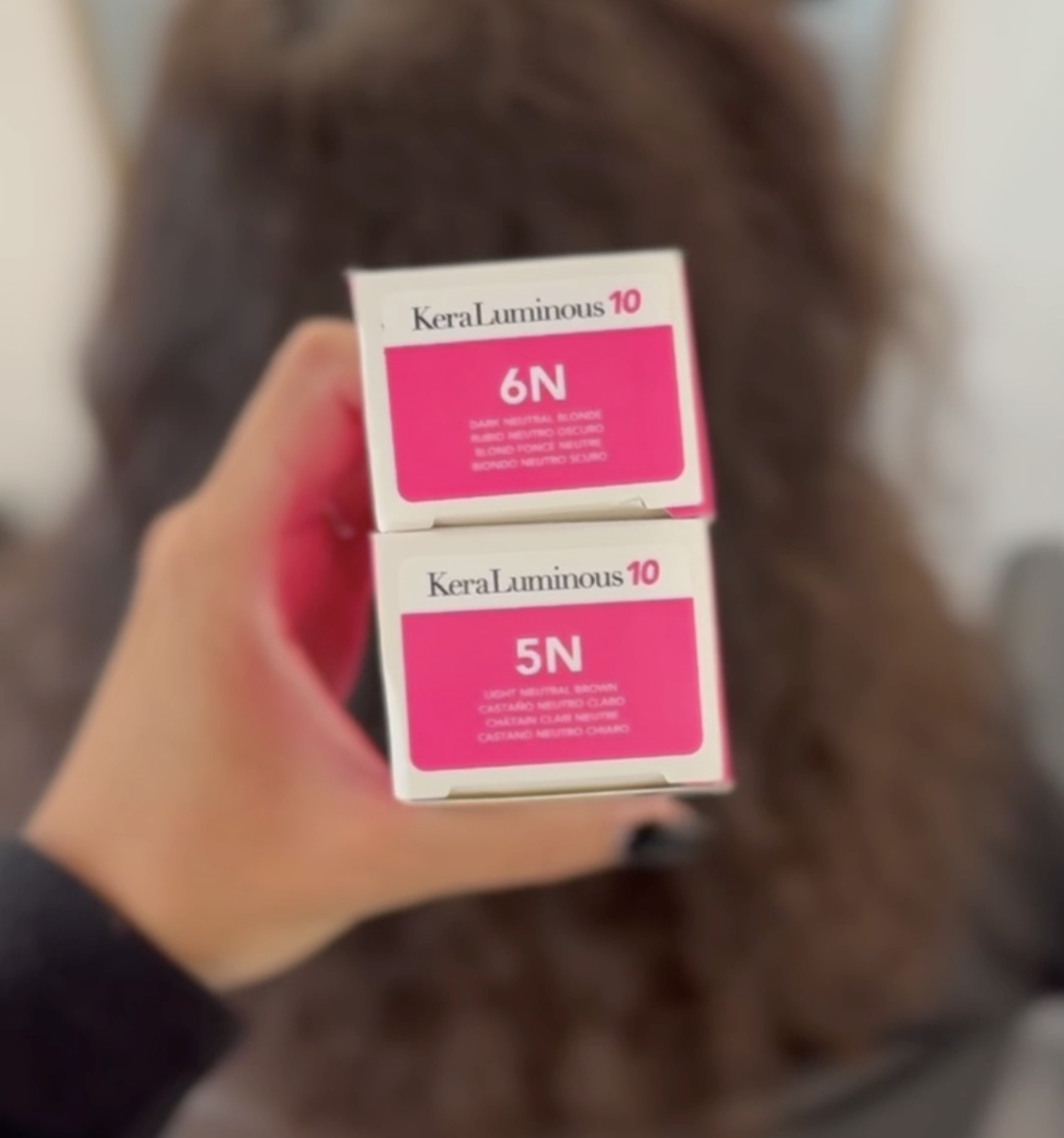 Hand holding two Keratin Complex KeraLuminous 10 hair color boxes labeled 6N Dark Neutral Blonde and 5N Light Neutral Brown.