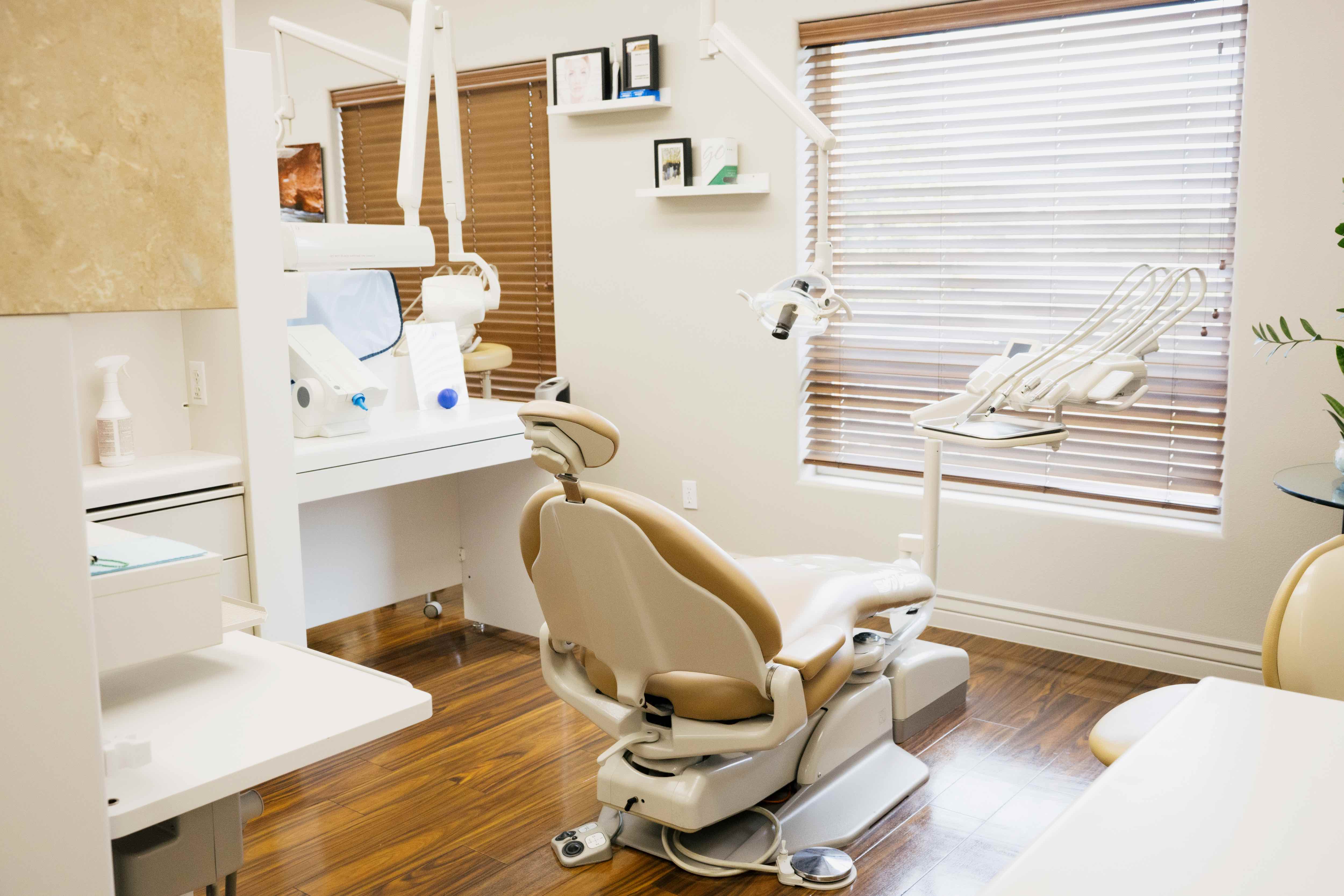 Bright dental office with beige dental chair, equipment, wooden floor, and window blinds.