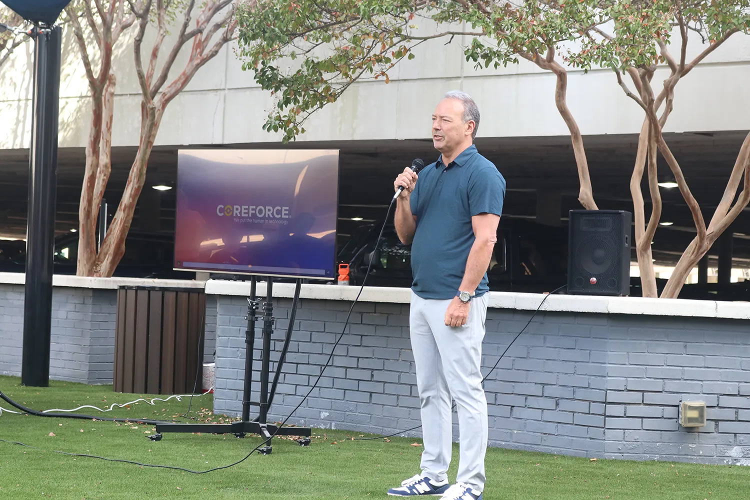 Man in a blue polo shirt and white pants speaking into a microphone outdoors next to a TV displaying Coreforce logo and a speaker.