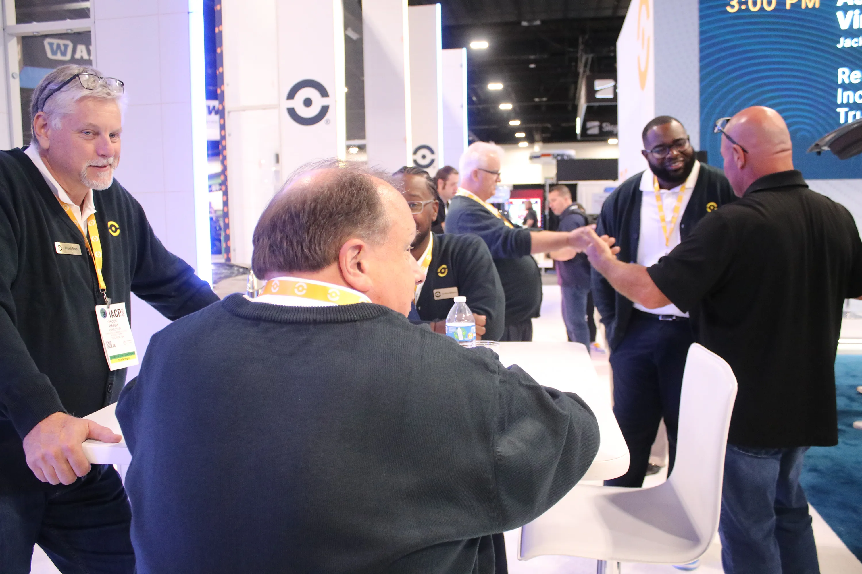 Group of six men wearing black sweaters with yellow lanyards gathered around a white table at an indoor event.