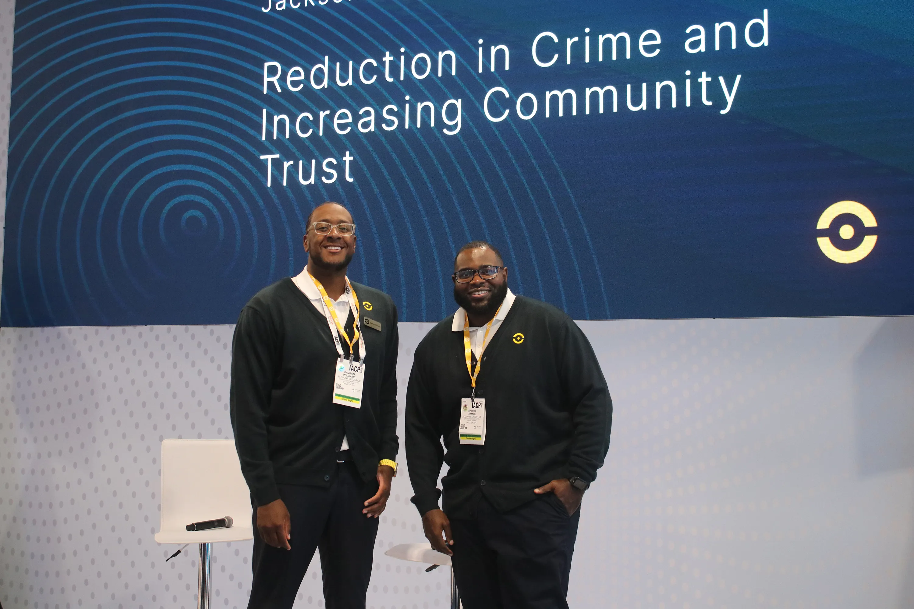 Two men wearing black cardigans with yellow lanyards smiling and standing in front of a screen displaying the text 'Reduction in Crime and Increasing Community Trust.'
