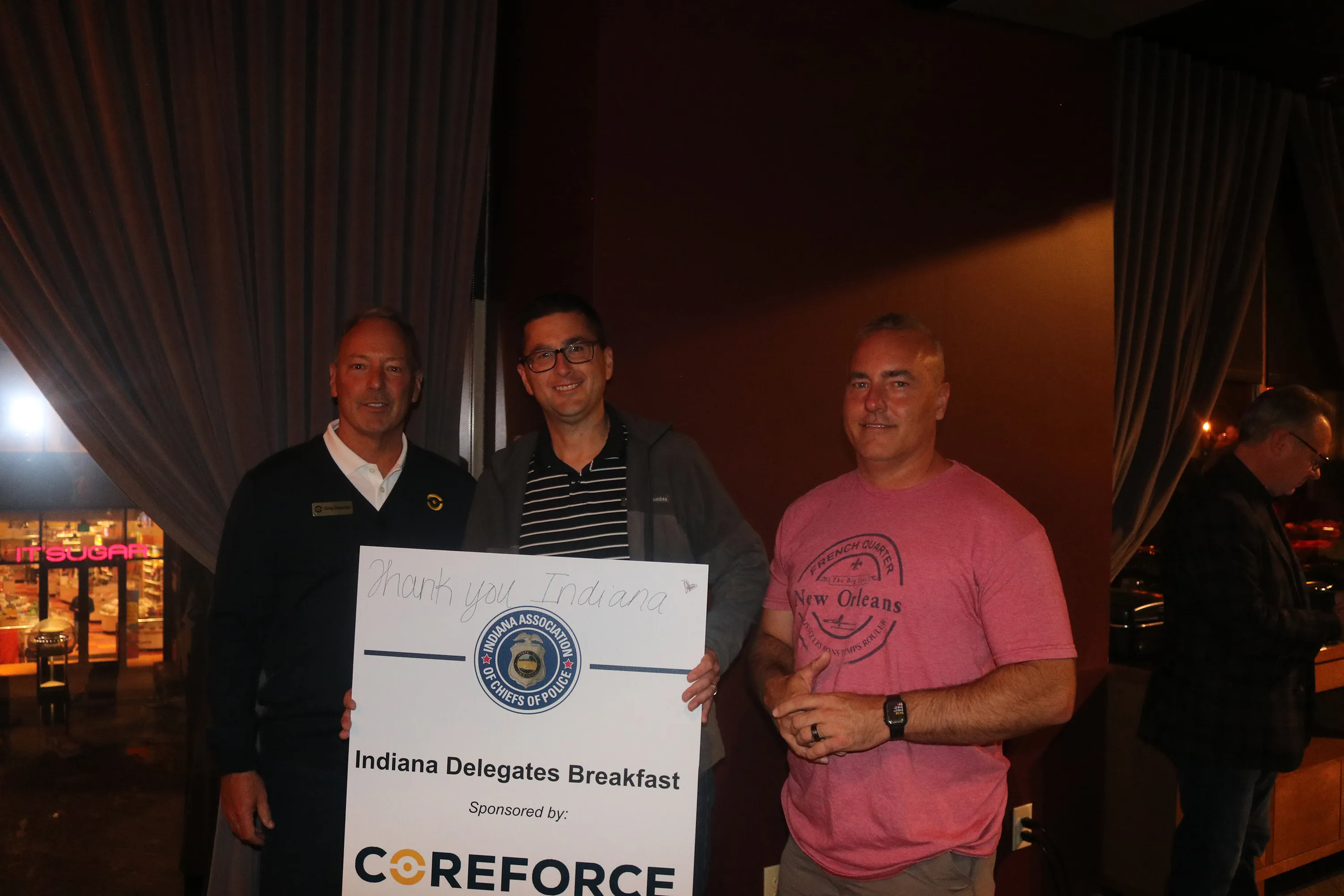Three men standing indoors, two holding a sign that reads 'Indiana Delegates Breakfast Sponsored by CoreForce' with the Indiana Association of Chiefs of Police logo.