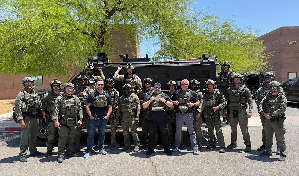 Group of armed police officers in tactical gear posing in front of a black SWAT vehicle under a tree on a sunny day.