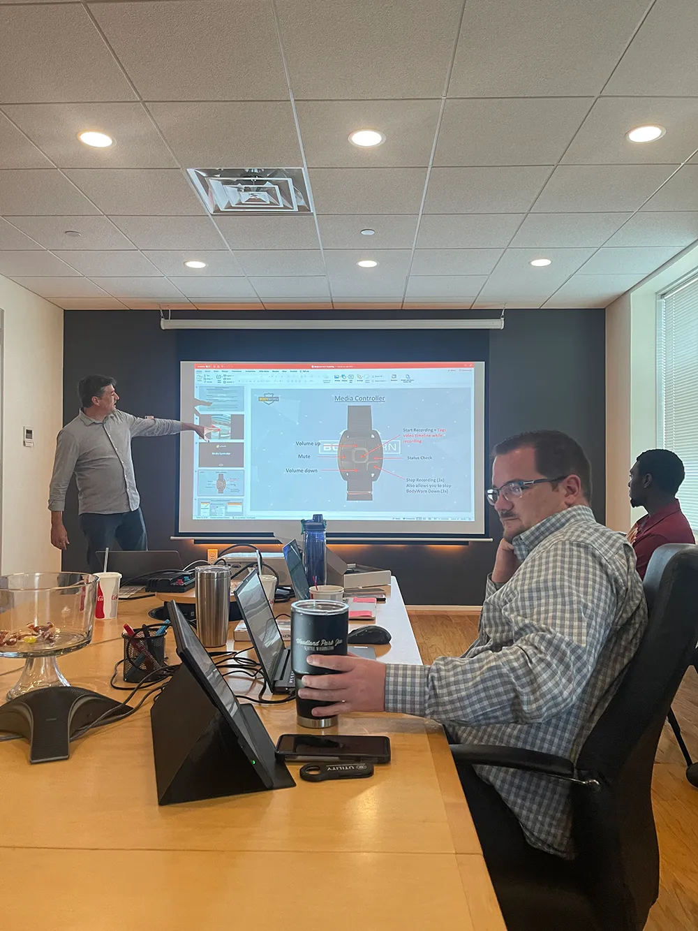 Man standing and pointing at a projection screen during a meeting, with two men seated at a table working on laptops and holding drinks.