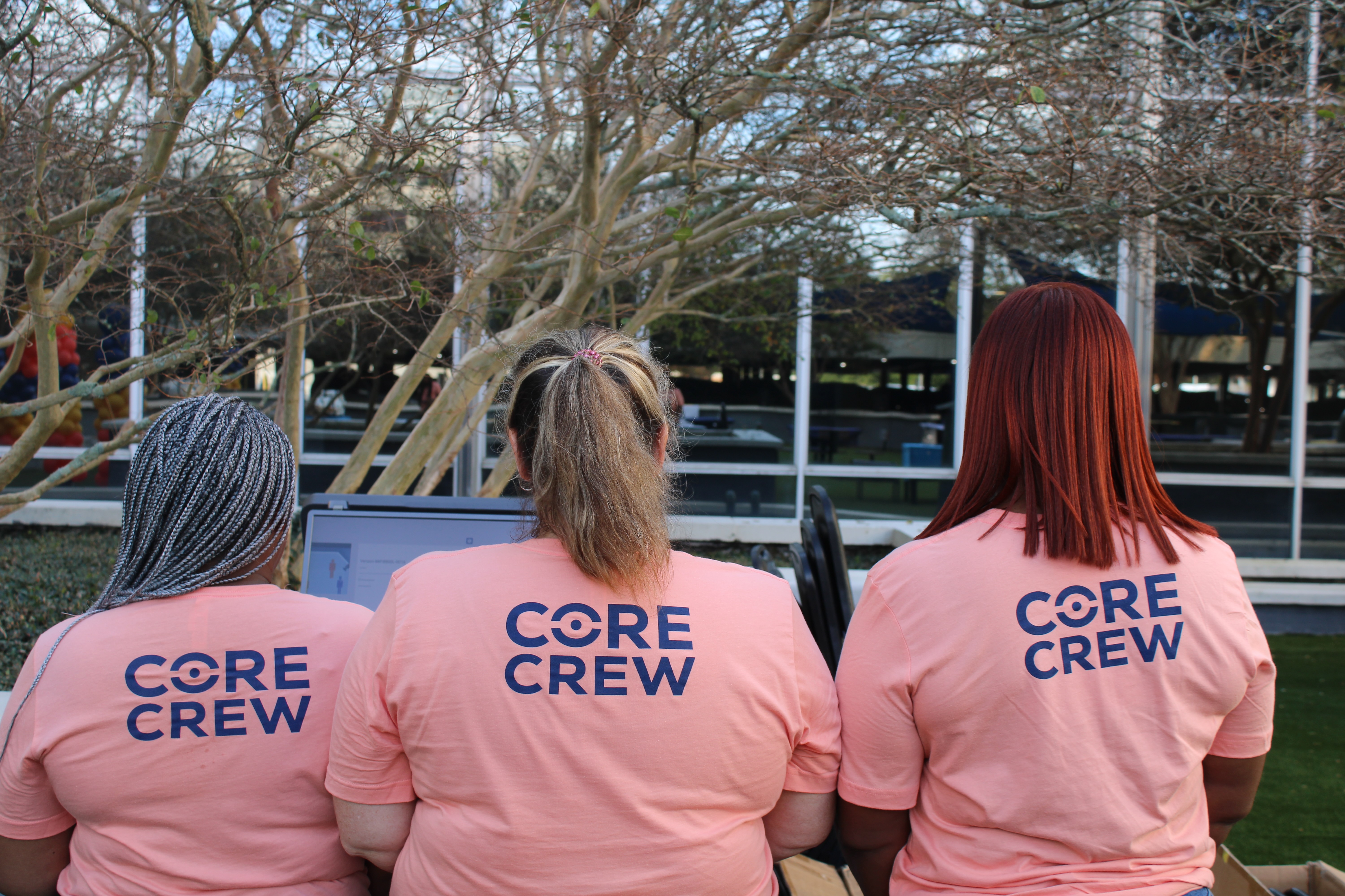 Three people with different hairstyles sitting side by side wearing matching pink 'CORE CREW' t-shirts.