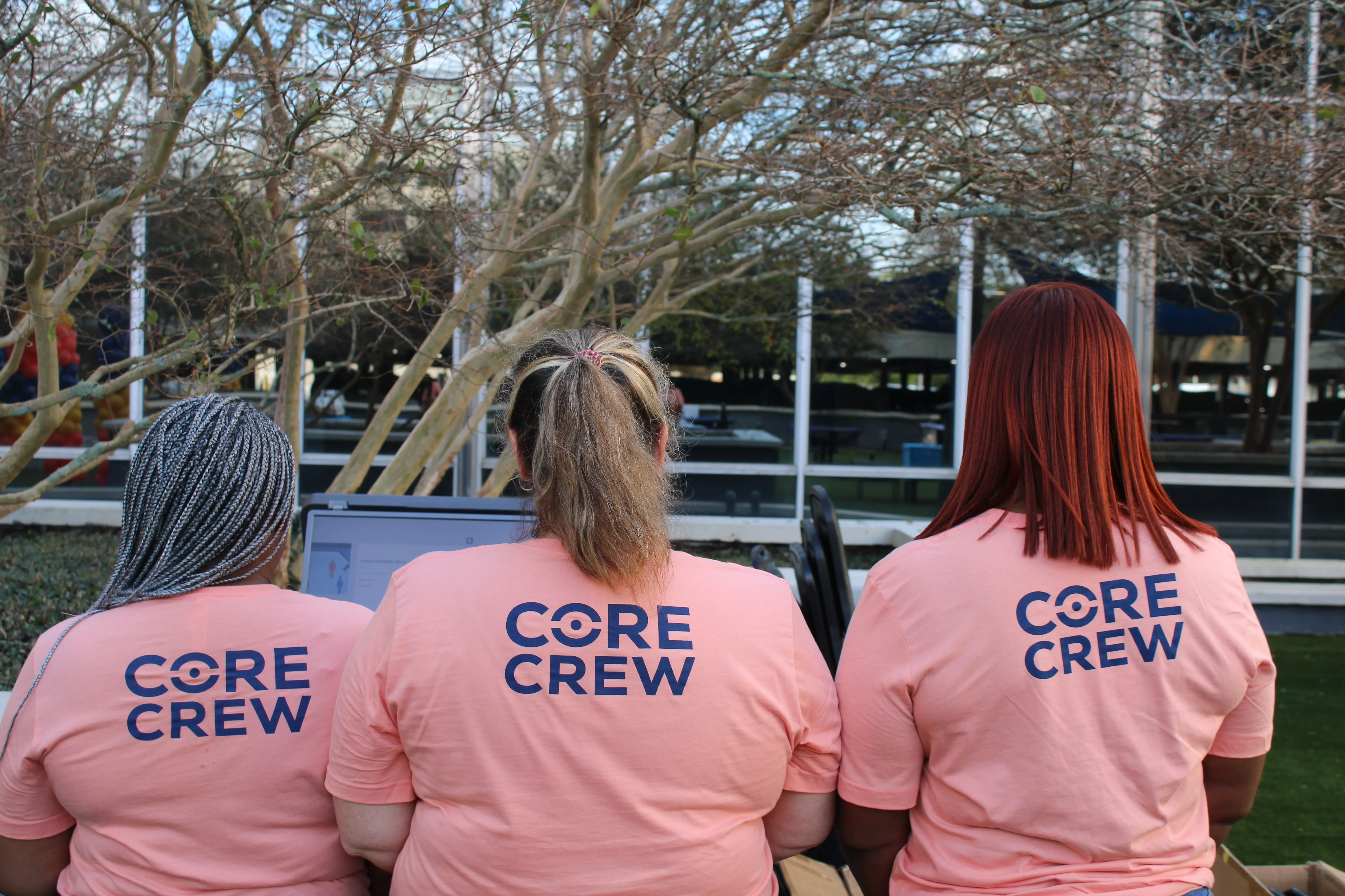 Three people with different hairstyles sitting side by side wearing matching pink 'CORE CREW' t-shirts.